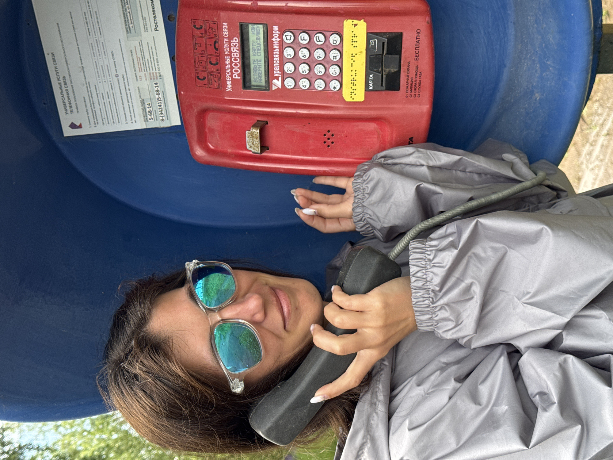 A young woman with brown hair and sunglasses holds a black telephone handset to her ear while leaning against a blue, outdoor payphone booth. She wears a grey hooded jacket. The payphone is red and has a keypad, a card slot, and Braille markings. A sign above the keypad reads "УНИВЕРСАЛЬНЫЕ УСЛУГИ СВЯЗИ РОСССЯЗЬ" (Universal communication services ROSSYAZ). The background consists of greenery and a bright sky, suggesting an outdoor setting. There is no specific indication of the location being in the city of Вассята, Russia.