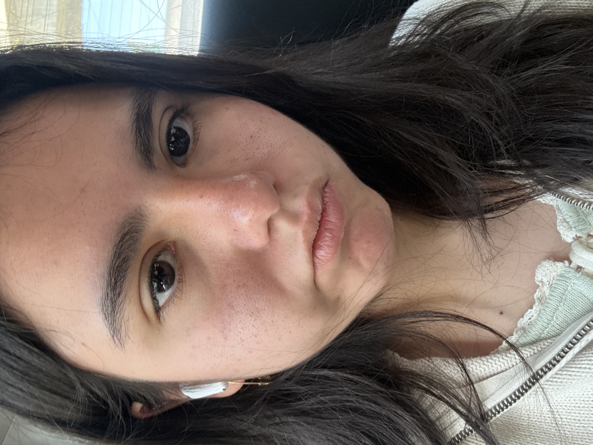 A selfie of a young woman with dark hair and brown eyes. She is wearing a white zip-up sweater and an earpiece in her left ear. Her skin tone is fair, and she has freckles on her face. A window is visible in the background. I cannot determine a location in Metepec, Mexico, based on the image.