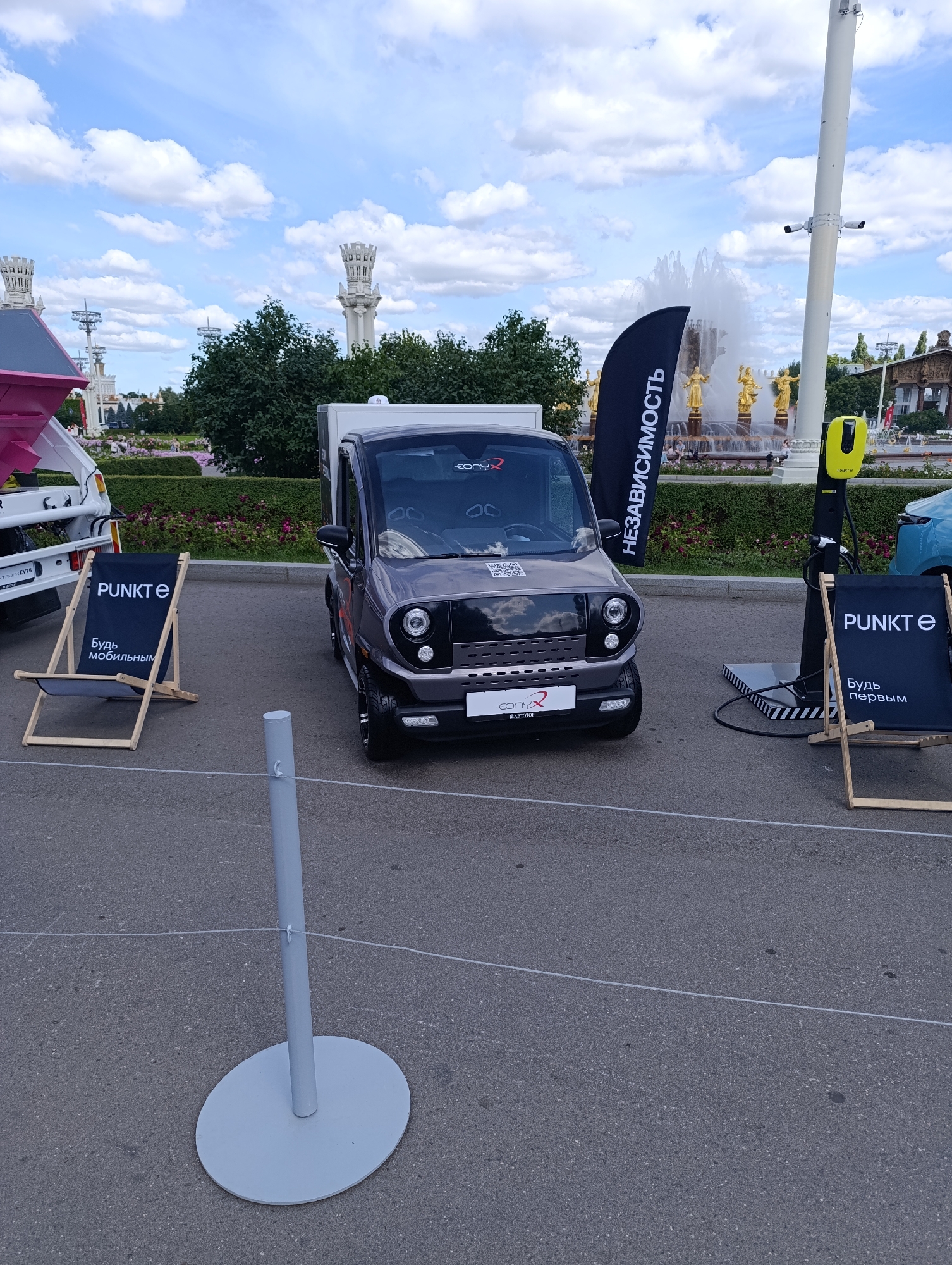 The image depicts an outdoor exhibition or event, likely at VDNKh (Exhibition of Achievements of National Economy) in Moscow, Russia, given the presence of iconic fountains and Soviet-era architecture visible in the background. In the foreground, a small, modern electric vehicle, possibly a delivery van or compact car, is parked. It has a dark gray body and a black front grille with circular headlights. A QR code is visible on the hood. To the left of the vehicle, a black deck chair with white lettering that reads "PUNKTE" and "Будь мобильным" (Be mobile) is set up. To the right of the vehicle, another similar deck chair with "PUNKTE" and "Будь первым" (Be first) printed on it is positioned next to an electric vehicle charging station, which is yellow and mounted on a stand with black and white stripes. A black banner with white Cyrillic text, "НЕЗАВИСИМОСТЬ" (Independence), is also visible, partially obscuring the fountain in the background. The overall scene is set on an asphalt surface with a light-colored stanchion and thin white wire in the lower portion of the frame, suggesting a cordoned-off area or exhibit space. The background features lush green trees, a large ornate fountain with golden statues, and distinctive architectural structures characteristic of VDNKh. The sky is blue with scattered white clouds. No people are clearly visible in the immediate vicinity of the vehicles or exhibits.
