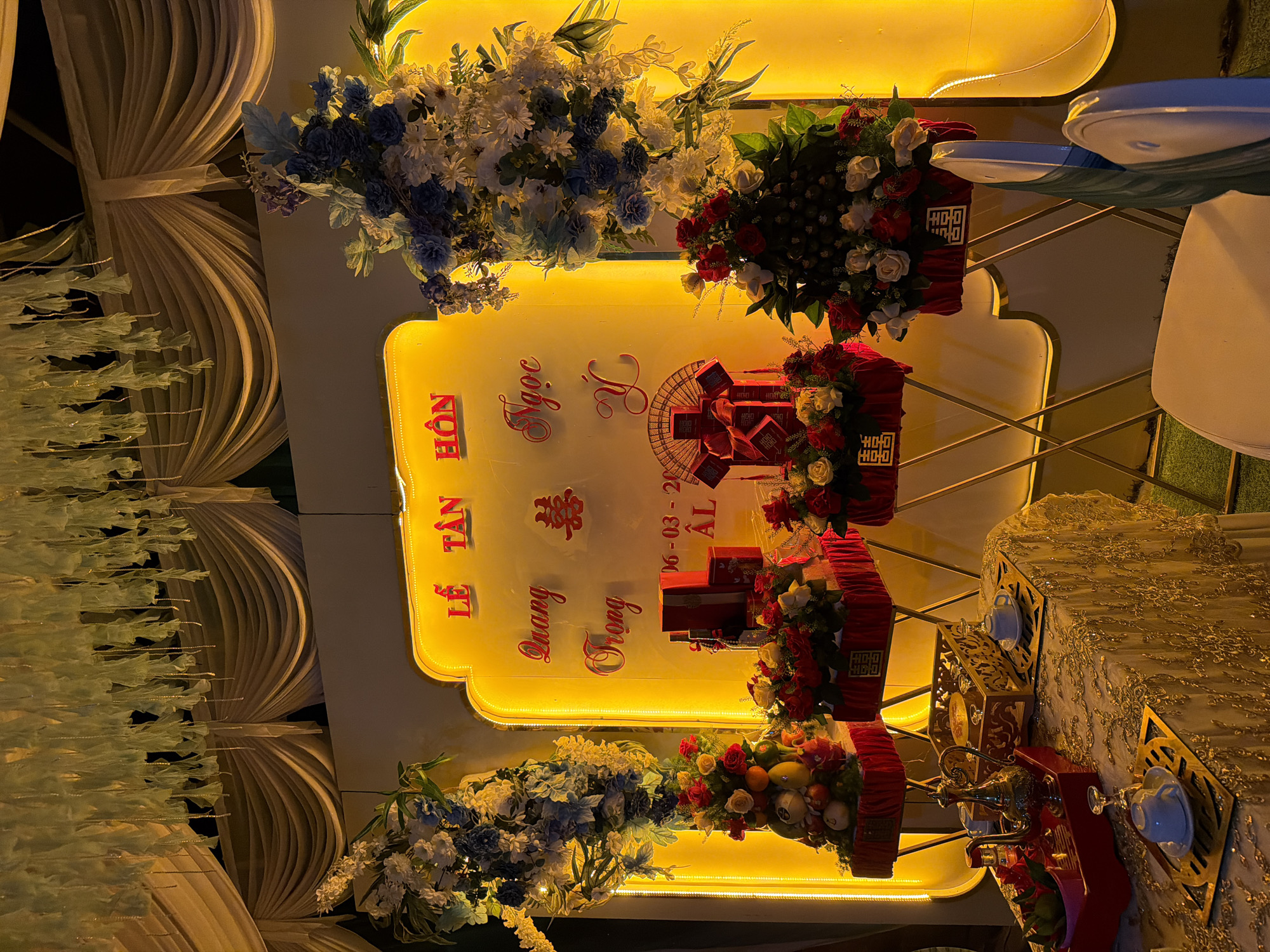 This image captures an elaborately decorated setting for a wedding or engagement ceremony, indicated by the prominent Vietnamese text "LỄ TÂN HÔN" (Wedding Ceremony/Reception). The scene is brightly illuminated with warm yellow light, suggesting an evening event or indoor setting.
At the center, a large, ornate yellow backdrop, shaped like a traditional plaque, glows warmly. Emblazoned on it in elegant red script are the names "Ngọc Y" and "Quang Trọng," likely the couple being celebrated. Above their names, the Chinese character for "double happiness" (囍) is displayed, and below them, the date "06-03-2023" is visible.
Flanking the central backdrop are large, decorative floral arrangements. To the left, a bouquet consists primarily of white and various shades of blue flowers with green foliage. To the right, another arrangement features vibrant red and white roses mixed with dark green leaves.
Below these displays, on tables, are multiple red gift boxes or trays, many of which are adorned with the "double happiness" symbol in gold. Some trays also hold arrangements of fresh fruits, such as oranges and pomegranates, alongside smaller flower clusters.
To the right of the frame, a table is visible, draped with an elegant white or cream tablecloth featuring intricate gold patterns. On this table, white teacups and saucers are placed within decorative gold stands, accompanied by what appears to be a golden teapot or urn. On the far left, draped light-colored fabrics or curtains form part of the background decor, contributing to the formal and celebratory atmosphere. The overall impression is one of a festive, carefully arranged cultural event.