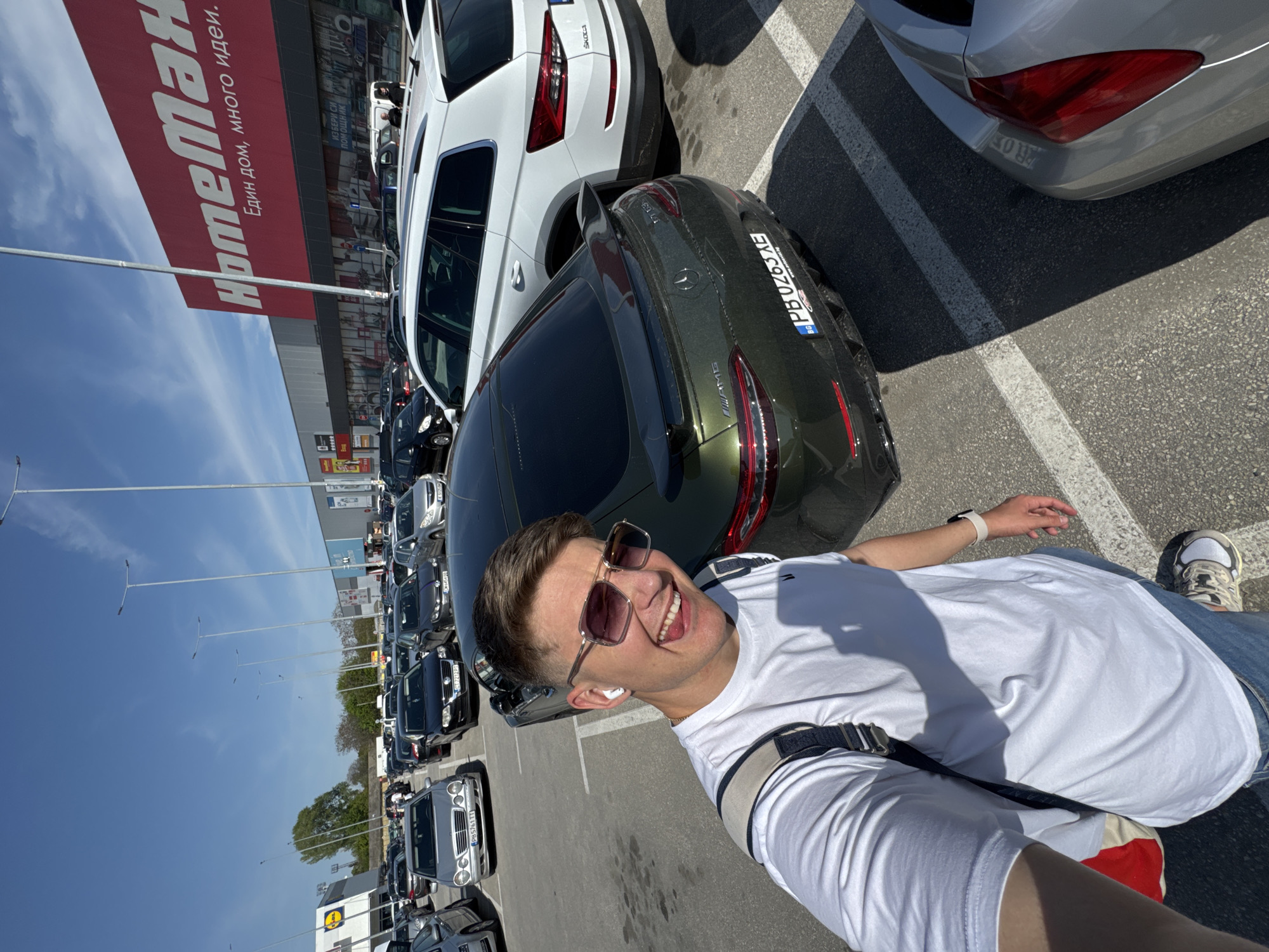 A young man with sunglasses and wireless earbuds is taking a selfie in a large outdoor parking lot under a clear, bright blue sky. He is smiling widely, showing a cheerful expression. He is wearing a white t-shirt, blue jeans, a white smartwatch, and a small black shoulder bag.
The parking lot is filled with numerous cars, indicating a busy shopping area. Behind the man is a dark green Mercedes-Benz AMG, prominently displaying a Bulgarian license plate "PB0263XE" (Plovdiv region). A white Skoda SUV is parked adjacent to the Mercedes. Further in the background, a white car with license plate "PB5761TT" is also visible.
In the background, large retail buildings can be seen. A prominent red sign reads "HomeMaN" (Homeman) with the tagline "Един дом, много идеи." (One home, many ideas.) in Cyrillic script. Another building sign, partially visible, appears to be for "LIDL". Other text on buildings includes "ИЗБЕРИ СИ ПОМОЩНИК" (Choose a helper). The bright sun suggests it is daytime. Based on the license plates and store names, the location is Plovdiv, Bulgaria.