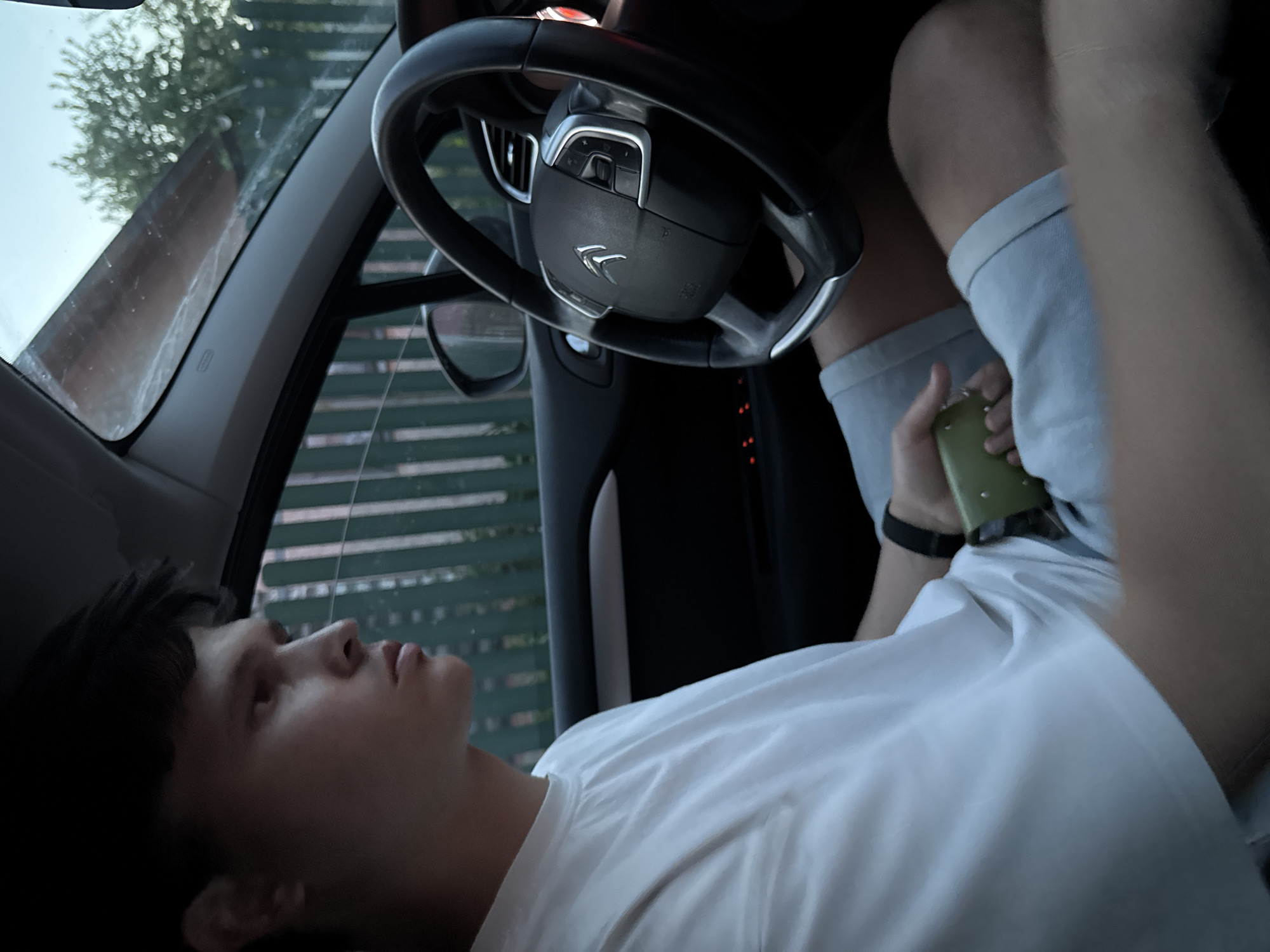 A person is reclining in the driver's seat of a car, appearing to look upwards and slightly to the left. They are wearing a white t-shirt and light-colored shorts. Their left arm is raised, holding a green object that may be a wallet or phone. A black smartwatch is visible on their left wrist. The car's steering wheel, with a Citroen logo, is in the foreground. The background through the windshield shows a blurry exterior with trees and a building. It is not possible to confirm the location is Mayak, Russia, from the given image.