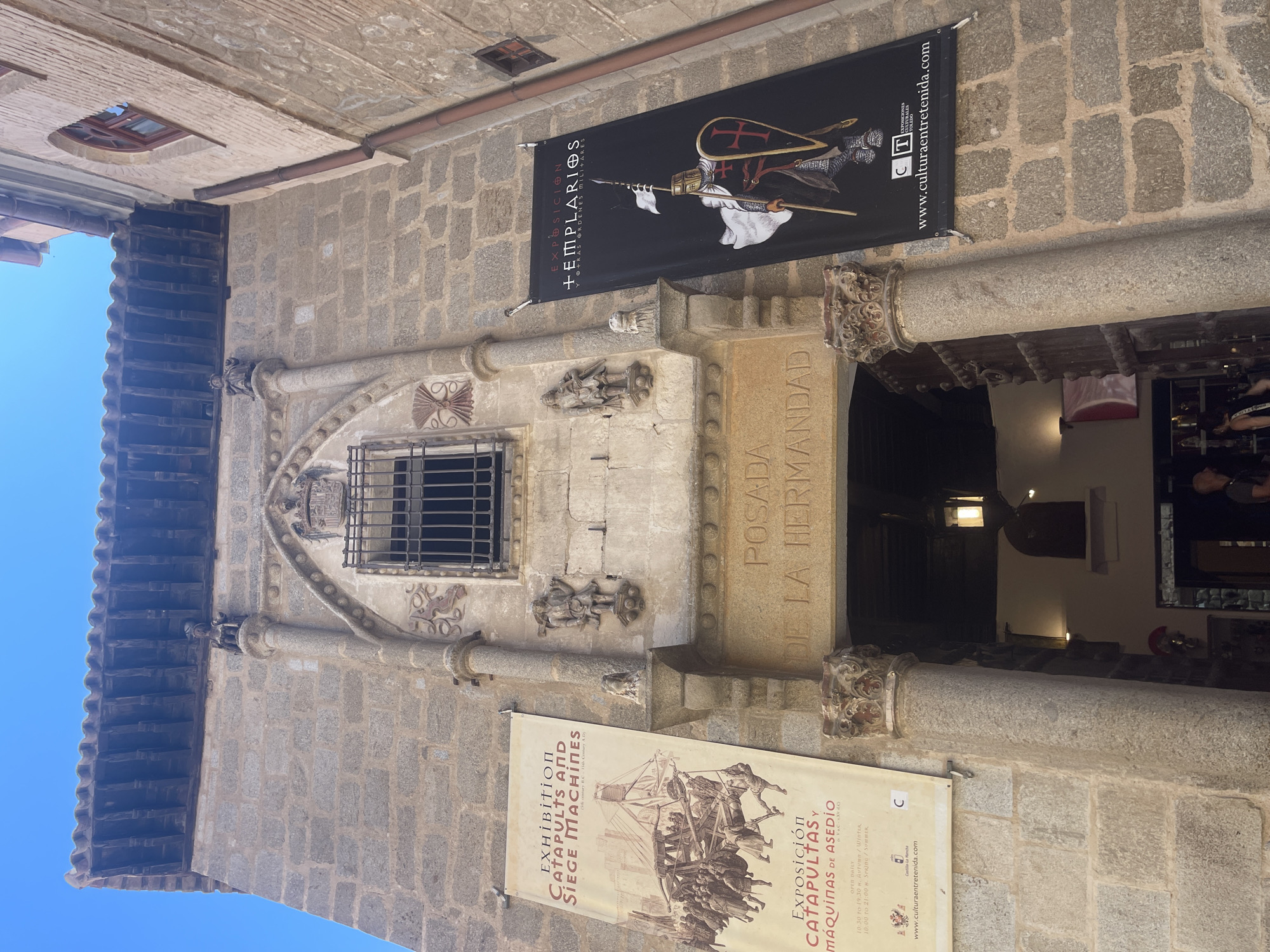 The image shows the entrance to the "Posada de la Hermandad" in Toledo, Spain. The building is constructed of stone, with a red-tiled roof. Two banners hang on either side of the entrance. The left banner advertises an exhibition of catapults and siege machines, and the right banner advertises an exhibition about the Templars. The entrance itself is a large stone archway with two large wooden doors. Above the archway, there is a small window with a wrought-iron grille. Two small stone statues are visible on either side of the window, and two larger statues are positioned on columns flanking the entrance. A wrought-iron lantern hangs inside the entrance. The sky is clear and blue. The scene is a street view of an historic building.