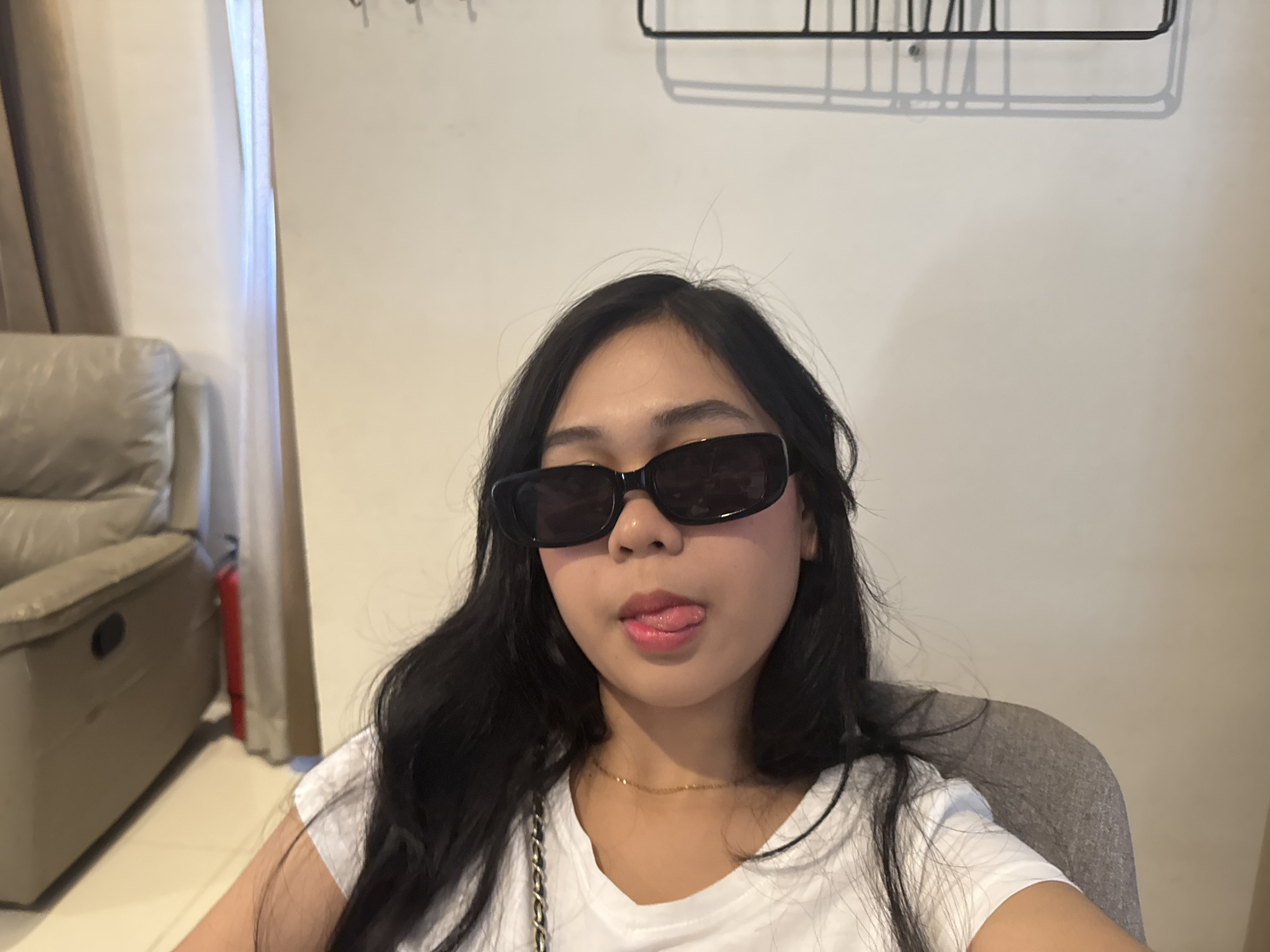 The image shows a young woman with long, dark hair wearing black rectangular sunglasses and a white t-shirt. She is sticking her tongue out slightly. A thin gold necklace is visible around her neck. She is sitting in a grey chair in what appears to be a living room, with a beige recliner sofa visible in the background to the left, and a curtain behind it. A black wire shelf hangs on the wall behind her. The overall lighting suggests an indoor setting, possibly during the day. The image quality is moderately clear. There is no specific indication in the image to place it in Davao City, Philippines.