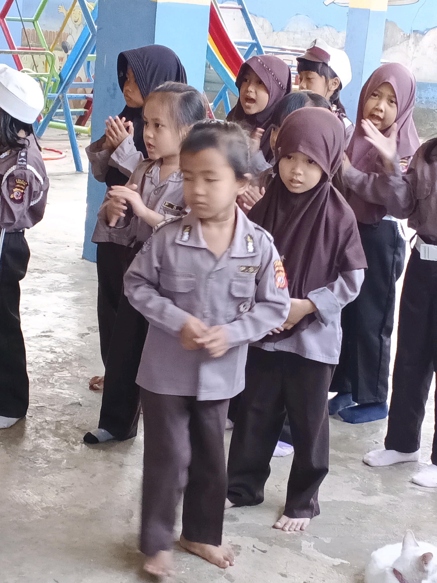 In Warungkiara, Indonesia, a group of young children are gathered outdoors, possibly at a playground or school. The children are dressed in what appears to be police uniforms, consisting of gray shirts with badges and dark pants. Some of the children are wearing headscarves in various colors, including black and maroon. One child in the foreground is looking down with their hands clasped. Other children in the background are clapping or looking towards the front. The ground is a textured concrete surface. A colorful playground structure is visible in the background, suggesting an outdoor recreational or educational setting. The overall scene depicts a group of children engaged in an activity, likely part of a school event or ceremony.