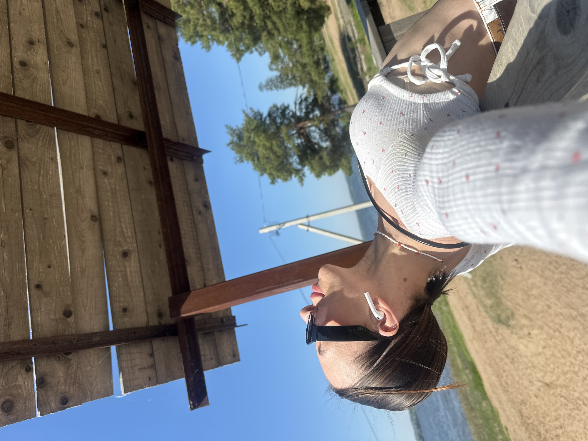 A low-angle, medium shot captures a young woman wearing sunglasses and a white top with a red floral pattern. She has an earbud in her ear and is wearing a beaded necklace. She is positioned against a bright blue sky. A weathered wooden structure is visible on the left, contrasting with the clear sky. In the background, there's a glimpse of a landscape featuring trees and water, suggesting an outdoor location. Without more information, it is not possible to confirm a location in Votkinsk, Russia.