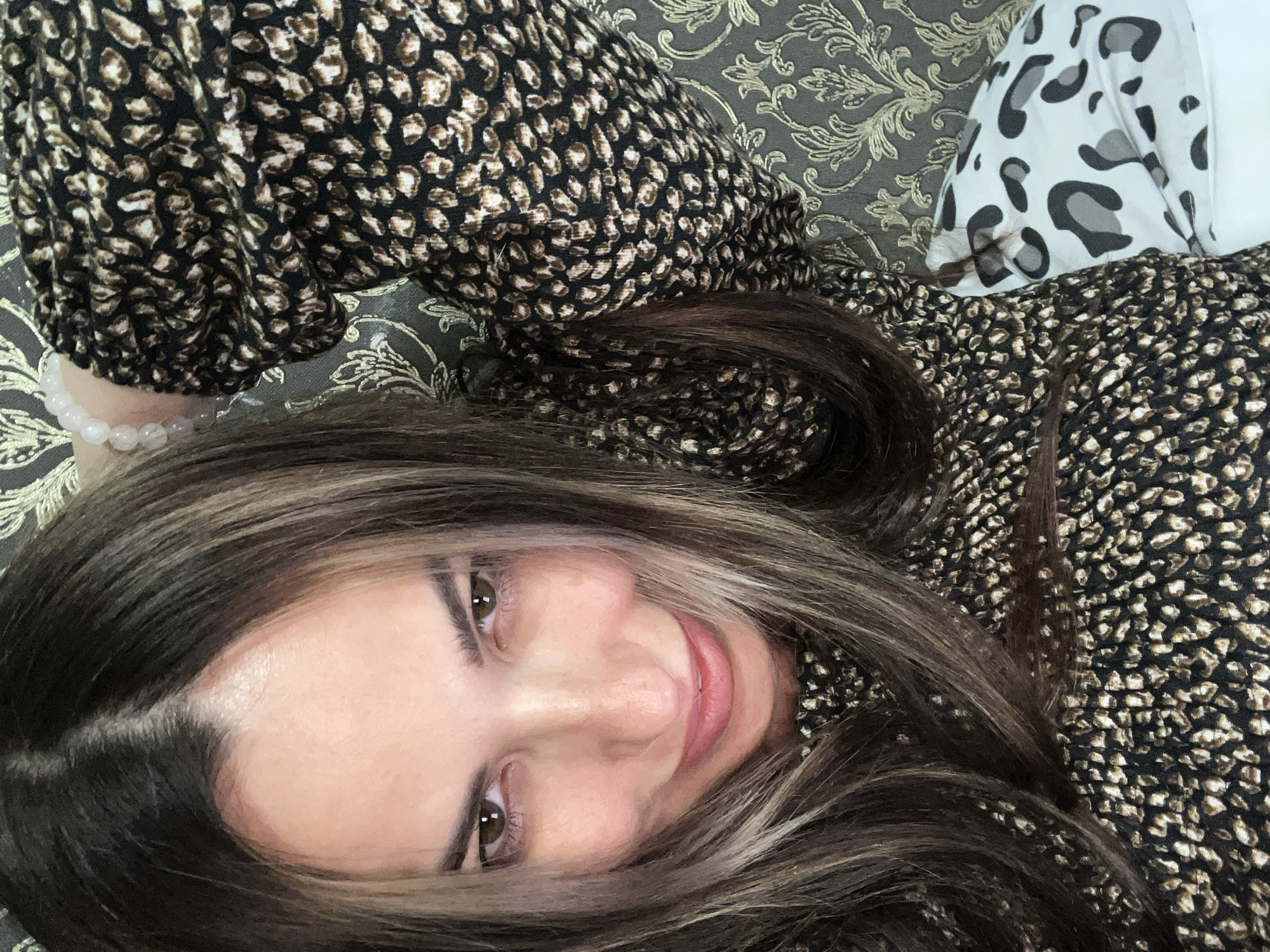 A woman with long, dark, highlighted hair and brown eyes is captured in a close-up, high-angle selfie. She is wearing a black dress with a subtle leopard print. Her left arm is bent upwards behind her head, and she is wearing a bracelet with light pink beads on her wrist. The background consists of a textured grey wallpaper with a gold floral pattern, and a white pillow with a grey and black leopard print is visible in the lower right portion of the frame. The lighting appears to be natural, coming from the front.
