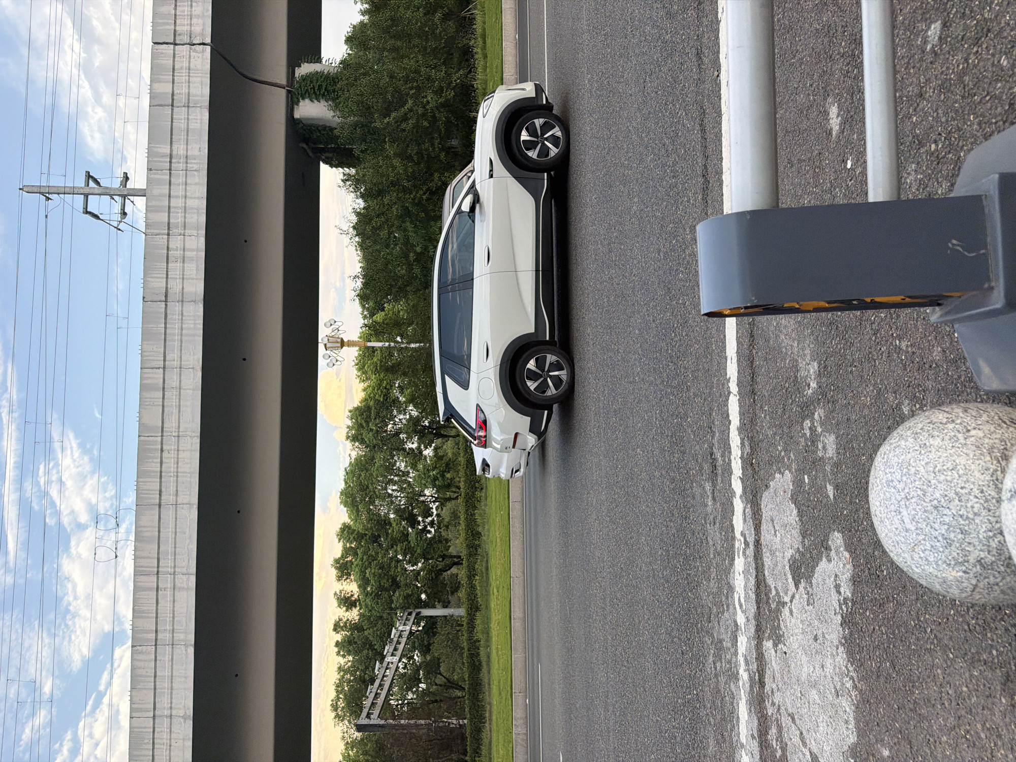 The image shows a white SUV on a road in Jiangjin District, Chongqing, China. The car is in motion, and another white vehicle is visible behind it. The road appears to be a multi-lane highway, with overhead infrastructure indicating an elevated structure, possibly a bridge or overpass, for transportation. Trees and greenery line the side of the road, and power lines are visible against a cloudy sky. The foreground shows a section of the road with a white line marking, and a gray barrier with orange reflective elements. The overall scene depicts a typical urban or suburban roadside environment.