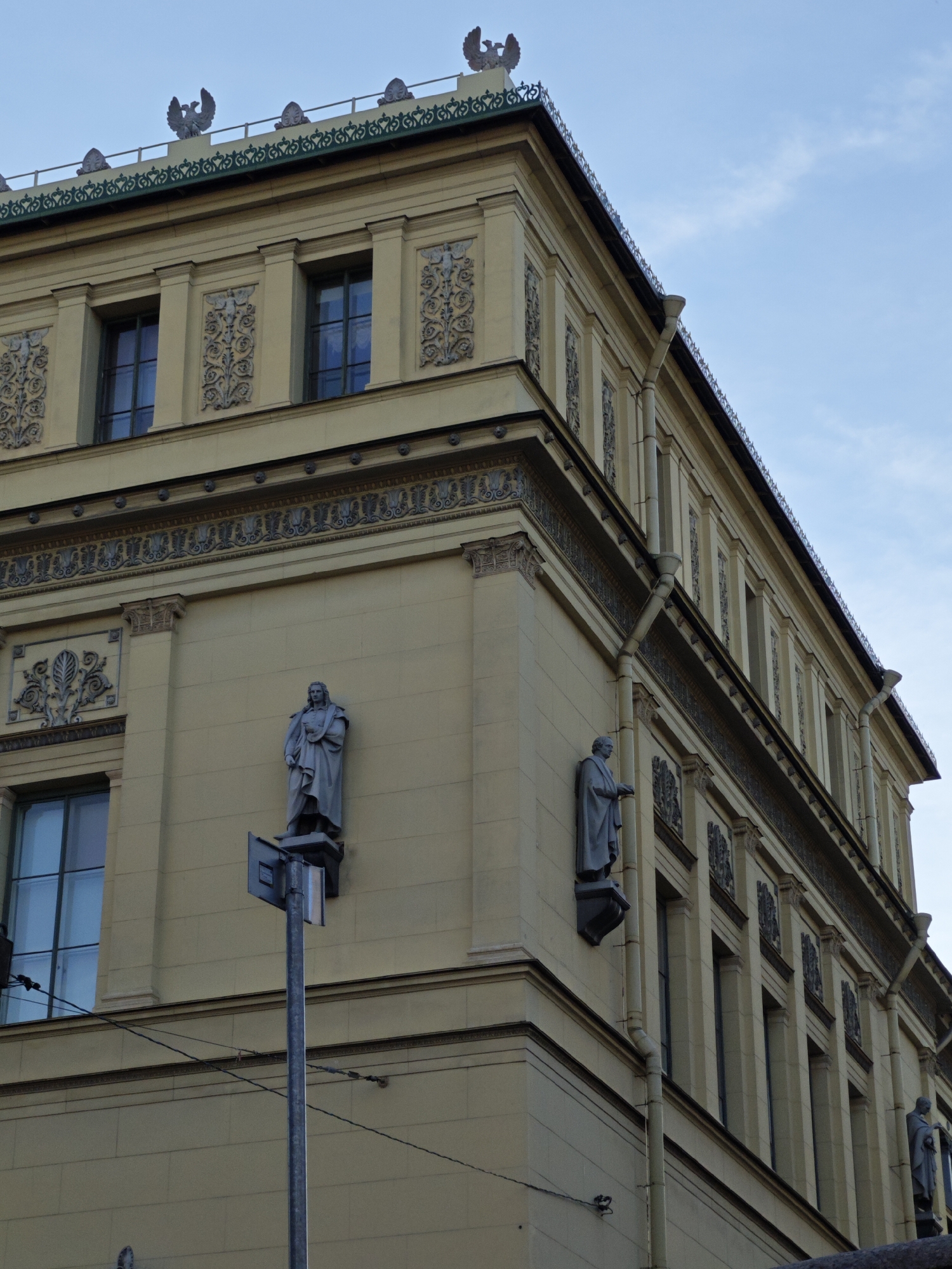 The image shows the exterior of a pale yellow building with a light blue sky overhead. The building has a classical architectural style, with decorative friezes and molding. Several statues of figures in draped robes are mounted on the facade, some of which appear to be eagles. There are tall, rectangular windows with multiple panes. The building has visible gutters and downspouts. The scene is a low-angle shot looking up at the building from street level.