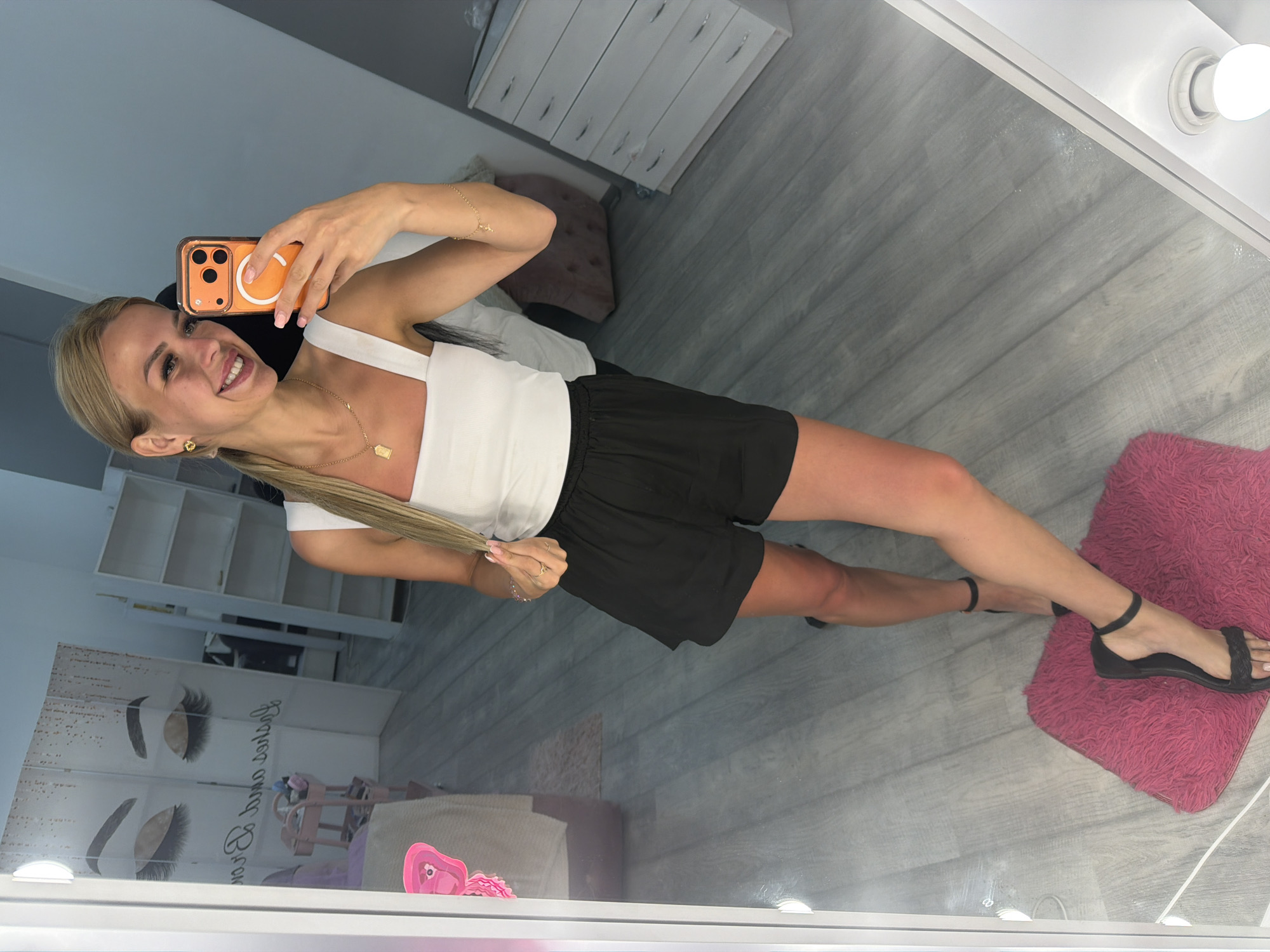 A woman is taking a selfie in a mirror. She is smiling and holding an orange phone in her right hand. She is wearing a white tank top and black shorts. She has blonde hair and is wearing gold jewelry. In the background, there are shelves and a dresser. The scene appears to be an indoor setting, possibly a salon given the decor. The lighting suggests it is daytime. The woman appears happy and confident. There is no visible text in the image.