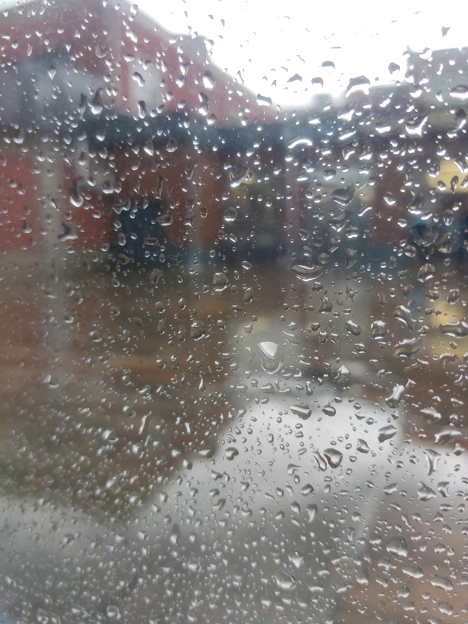 The image is a close-up view of raindrops on a window pane. Through the distorted glass, a blurry, muted cityscape is visible. The architecture appears to be residential or commercial, with hints of brick buildings and possibly other structures in the background. The sky is overcast, suggesting a rainy or gloomy day. The scene's location in Noginsk, Russia, is not visually evident from the image itself. There are no people, animals, or specific actions depicted. The focus is entirely on the texture and visual effect of the water droplets on the glass and the indistinct background.