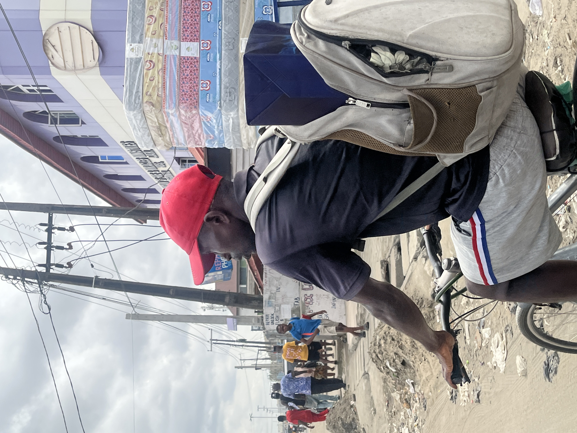 A man wearing a red cap and dark blue shirt is riding a bicycle laden with goods down a street. He has a large backpack, and rolls of mattresses are stacked precariously above. He looks downwards. The bicycle tires are visible.
The street scene includes pedestrians of varying ethnicities walking and standing. There are power lines overhead. Buildings are in the background. Trash is present along the roadside. The location appears to be Ado, Nigeria, indicated by signage visible in the background.