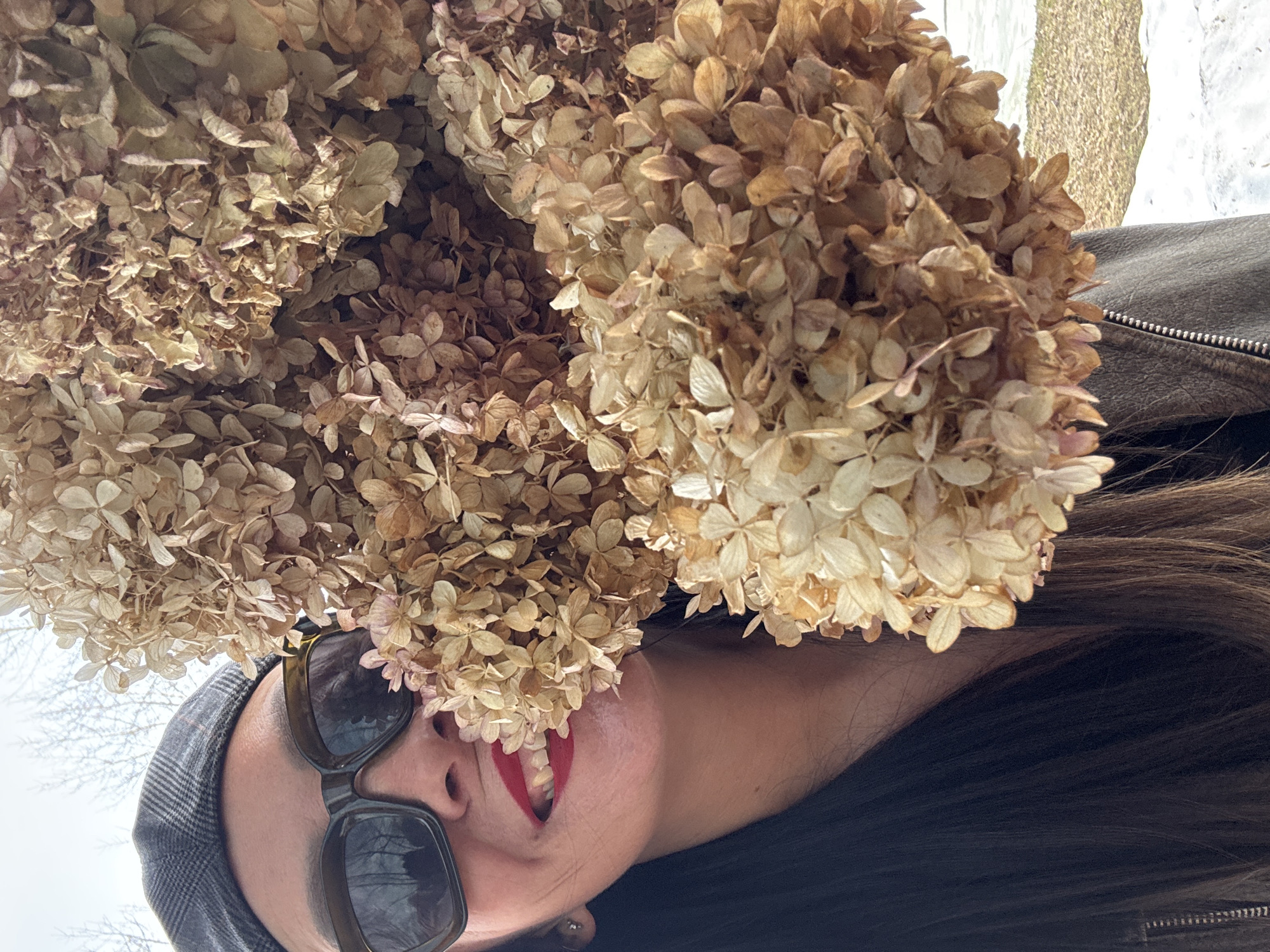 A woman wearing a gray cap and sunglasses, red lipstick, and a black leather jacket is holding a large bunch of dried hydrangeas. The location appears to be outdoors, possibly in a park or garden, based on the presence of trees and snowy ground in the background. The image does not provide enough information to definitively confirm the location as Chaikovsky, Russia.