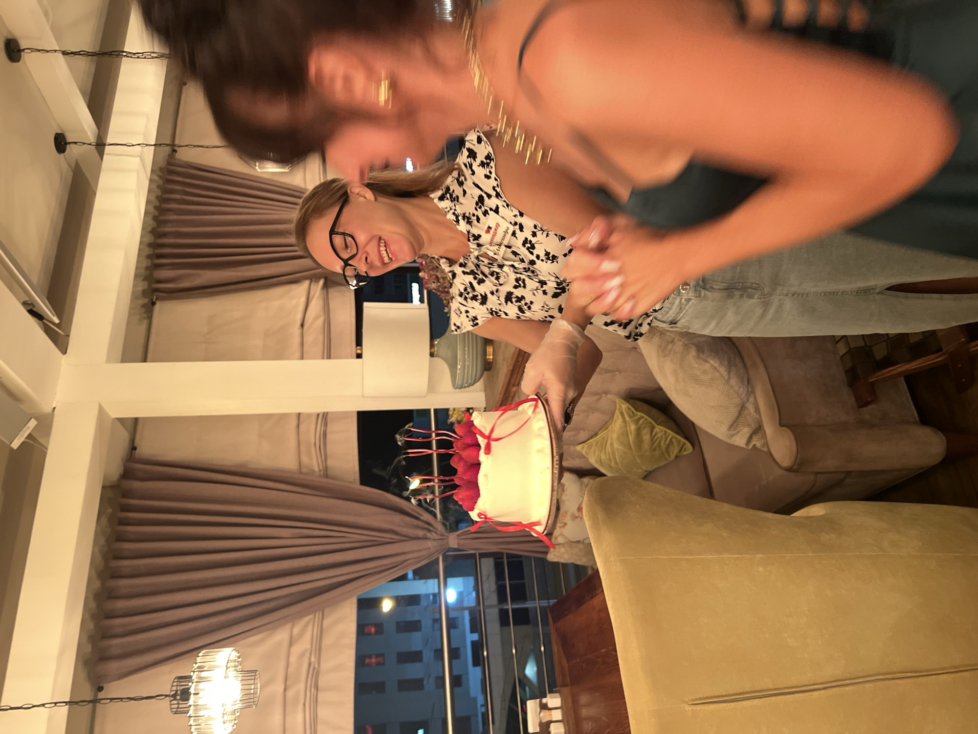 The image depicts a woman in a black and white floral print blouse and a light blue denim skirt, wearing black-rimmed glasses, holding a small white cake with red ribbons. The cake has several lit candles on top, surrounded by what appear to be red strawberries. The woman is smiling broadly and looking towards another person, whose face is out of focus and blurred. Her hands are gloved. The background shows a dimly lit interior with large windows offering a view of city buildings at night. The scene appears to be indoors, possibly a restaurant or a private residence. The presence of a cake with candles suggests a celebration, likely a birthday. The woman's attire and the overall ambiance suggest a casual yet festive occasion. There is no information in the image to suggest this location is in Novosibirsk, Russia.