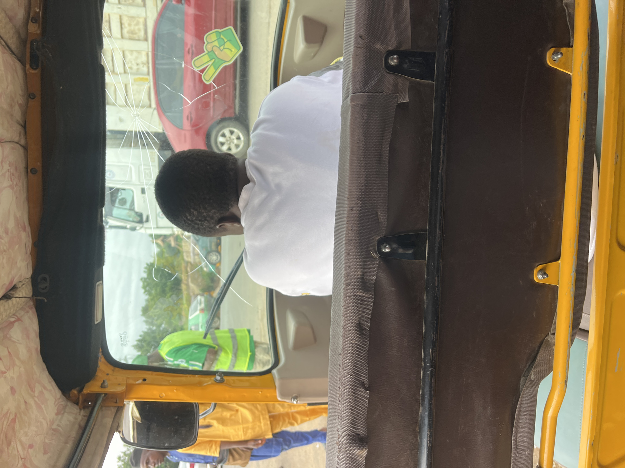 The image captures an interior view from a yellow tricycle, commonly known as a keke NAPEP, looking forward through a cracked windshield in Karmajiji, Nigeria. The central subject is a person, seen from behind, with short, dark, curly hair, wearing a plain white t-shirt, occupying the driver's seat.
The windshield is notably damaged, displaying a prominent crack originating from a central impact point, with numerous lines radiating outwards across the glass. Affixed to the upper right corner of the windshield is a vibrant green and yellow sticker depicting a hand making a peace sign (V-gesture).
Through the cracked and somewhat reflective glass, a bright daytime scene is visible. Elements outside include a red car, a white truck, and another person dressed in a bright green and yellow reflective vest, suggesting an urban or semi-urban setting. The interior of the tricycle features brown fabric covering structural pillars and parts of the roof, complemented by a lighter, patterned fabric on the ceiling. A side mirror is visible on the left, reflecting more of the exterior. The image appears to be a candid shot taken from a passenger's perspective during daylight hours.