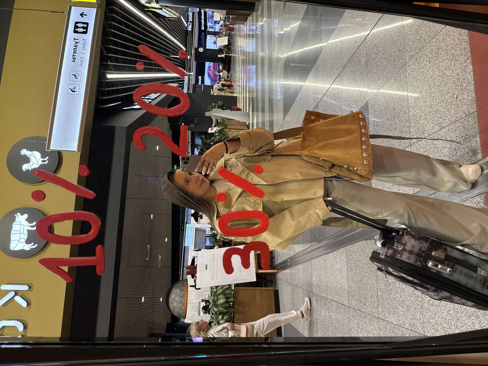 A person is taking a selfie in what appears to be a reflection in a store window, possibly in a shopping mall or an airport terminal in the city of Khimki, Russia. The store has signage in Russian, indicating its possible location in Russia. The reflections show various signs and directions, including one for toilets, a sign for 'Kope Cobon' in Russian, and a Mercedes-Benz shop. The person in the reflection is holding a phone up to take the picture. They are wearing a tan trench coat, light trousers, and a tan suede bag. They have luggage next to them. An older person is walking in the background. The foreground has lettering applied to the glass, apparently part of an advertisement.