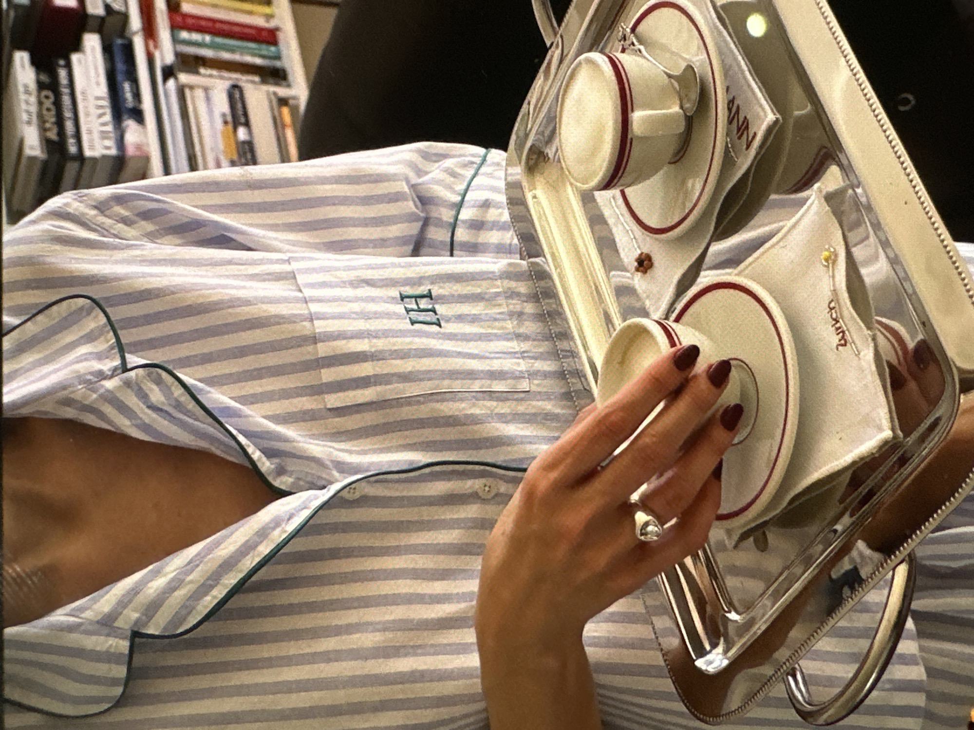 A person wearing striped pajamas is holding a silver tray with two coffee cups and saucers. The cups are white with a red stripe around the rim. The saucers have matching red stripes. There are also napkins on the tray, one of which reads "Zurich" and the other "ANN." The person has dark red nail polish and a ring on their finger. In the background, a bookshelf filled with books is visible. The location is not identifiable as Yerevan, Armenia from the provided image.
