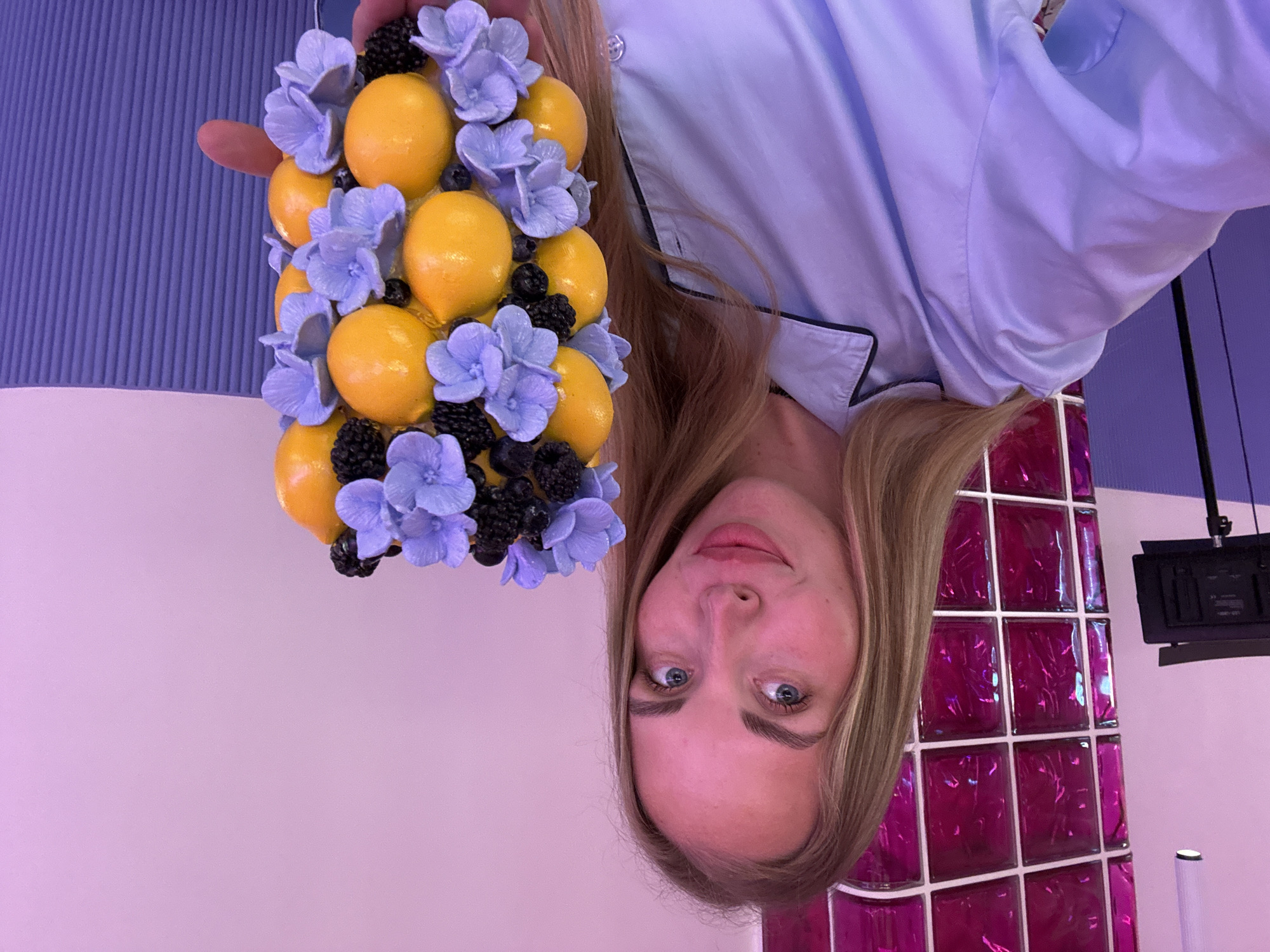 An indoor portrait captures a young woman posing while holding a decorative arrangement. The image is bright and colorful, set within a modern interior.
The subject is a young woman with long, straight blonde hair and striking blue eyes. She looks directly at the camera with a neutral, slightly smiling expression. She is wearing a light blue, long-sleeved shirt with dark piping details, reminiscent of a pajama top. Her left hand, adorned with light pink nail polish, holds a visually appealing bouquet.
This arrangement is composed of numerous artificial-looking, bright yellow lemons, intermingled with clusters of light purple flowers, possibly hydrangeas, and dark, spherical berries resembling blackberries. The elements are densely packed, forming a cohesive and vibrant piece.
The background of the scene features distinct sections. On the left, there is a textured wall or panel with vertical ribs in a muted lavender-purple hue. The central and lower background is a smooth, plain wall in a lighter shade of lavender-pink. To the right, a prominent feature is a vertical column of square, glossy tiles in a vibrant magenta or fuchsia color, which appear reflective or translucent, creating dynamic light patterns. Above these colorful tiles, a black electronic device, possibly a monitor or a light fixture, is mounted on the wall.
The woman is actively posing for the photograph, showcasing the unique fruit and flower arrangement. The overall aesthetic is characterized by a playful and modern use of color. The setting, identified as Sochi, Russia, suggests an interior space such as a contemporary cafe, hotel, or studio. No visible text is present in the image.