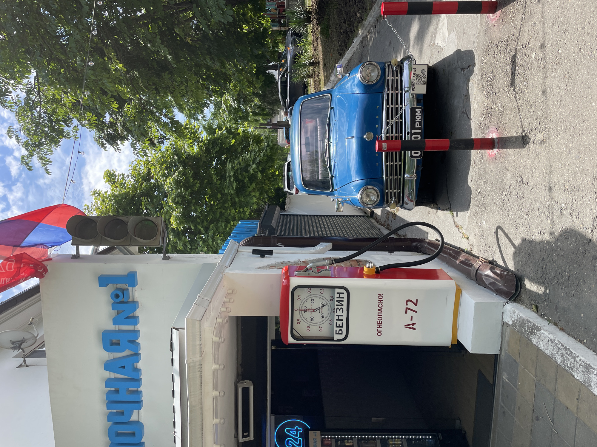 This image depicts a vintage blue car parked on a paved area in front of a building with a sign that reads "ПОЧНАЯ №1" in blue lettering. A vintage gas pump with a red top and white body stands to the left of the car. The gas pump has a gauge that reads "БЕНЗИН" and "ОГНЕОПАСНО!" below it. A license plate on the car reads "0 01 РЮМ" and has a QR code. Red and black bollards are placed in front of the car. The scene is framed by trees and a blue sky with clouds. The location is identified as a street in Anapa, Russia.