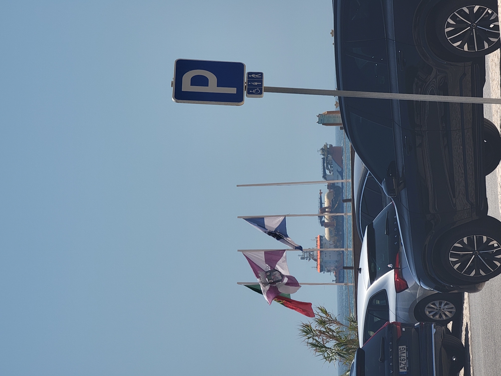 The image shows a parking lot with several cars in Quarteira, Portugal. In the foreground, a black SUV is parked on the right side of the frame, with its front wheel visible and a partial view of its side. To its left, a silver hatchback is parked, with its rear visible and a license plate that reads "AZ67VG". Further to the left, the rear of another black SUV is visible. In the background, three flags are flying on flagpoles. The flag on the left is red, green, and yellow. The middle flag is white and purple with a coat of arms. The flag on the right is blue and white. Beyond the flags, a large ship is visible on the water, with an orange and blue superstructure. The sky is clear and blue. A parking sign with a "P" symbol and symbols for disabled access and gender-neutral restrooms is visible on a pole in the middle right of the frame.