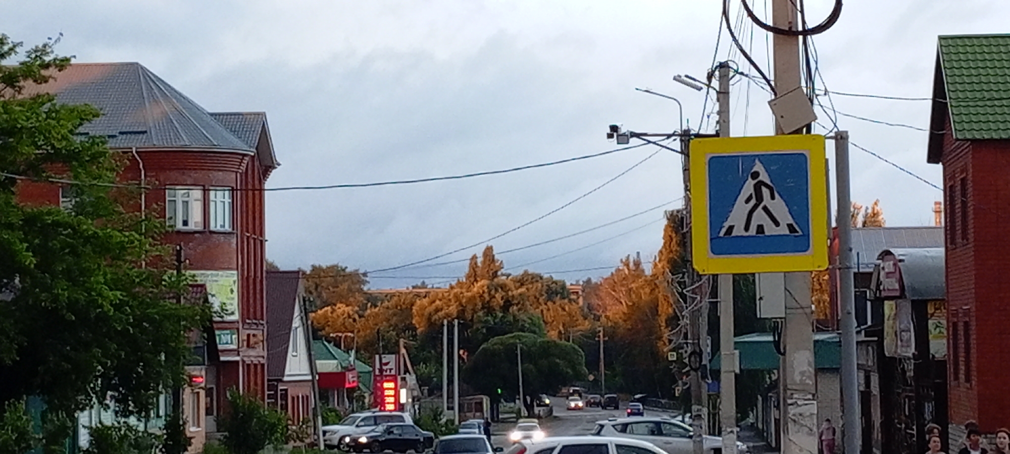 The image depicts a street scene in Kirsanov, Russia, under an overcast sky. On the left, a multi-story brick building with a dark tiled roof dominates the foreground. Adjacent to it, lush green trees partially obscure the building's lower facade, which features several windows and signage, including what appears to be a red traffic light. Further down the street, a gas station sign with red numerical displays is visible.
To the right of the street, a utility pole supports a pedestrian crossing sign and various wires. A dark red, two-story building with a green tiled roof is visible beyond the utility pole, with other buildings extending further down the street. The street itself is lined with trees that show autumnal colors, with foliage in shades of orange and yellow. Several vehicles, including cars and what appear to be vans, are present on the road, some in motion and others parked. A few individuals can be seen walking on the sidewalk. The overall atmosphere suggests a quiet street scene in a Russian town.