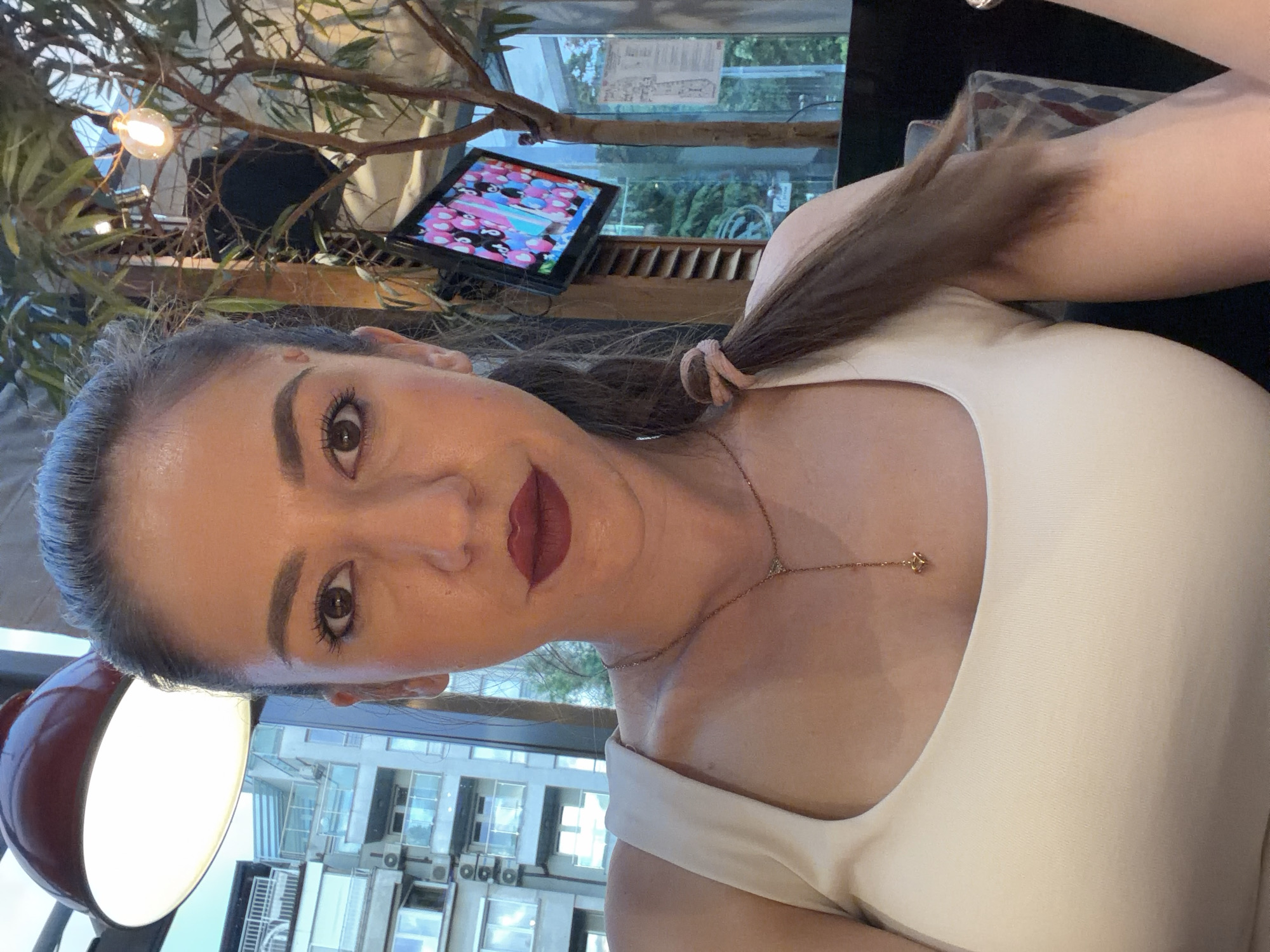 A female subject is depicted in a close-up, chest-up, frontal portrait, looking directly at the camera. She has dark hair, styled in a ponytail secured with a light pink scrunchie. Her makeup includes dark eyeliner, mascara, and dark red lipstick. She is wearing a plain, light-colored (likely cream or white) scoop-neck tank top. A delicate gold-toned necklace with a small, star-shaped pendant hangs around her neck, extending down her sternum.
The scene is set indoors, within what appears to be a cafe or restaurant in the city of Sofia, Bulgaria. A prominent red industrial-style pendant lamp with a bright white light source is visible in the upper left. To the right, decorative elements resembling bare tree branches with green foliage are present, along with an exposed filament light bulb. A flat-screen television is mounted on a vertical surface, displaying colorful animated content. Behind the subject and through large windows, an urban landscape is visible, featuring multi-story buildings with multiple balconies and exterior air conditioning units, typical of residential or commercial structures in a city environment. Greenery is also discernible in the background outside the windows. The lighting within the establishment combines bright artificial sources with natural light entering from the exterior.