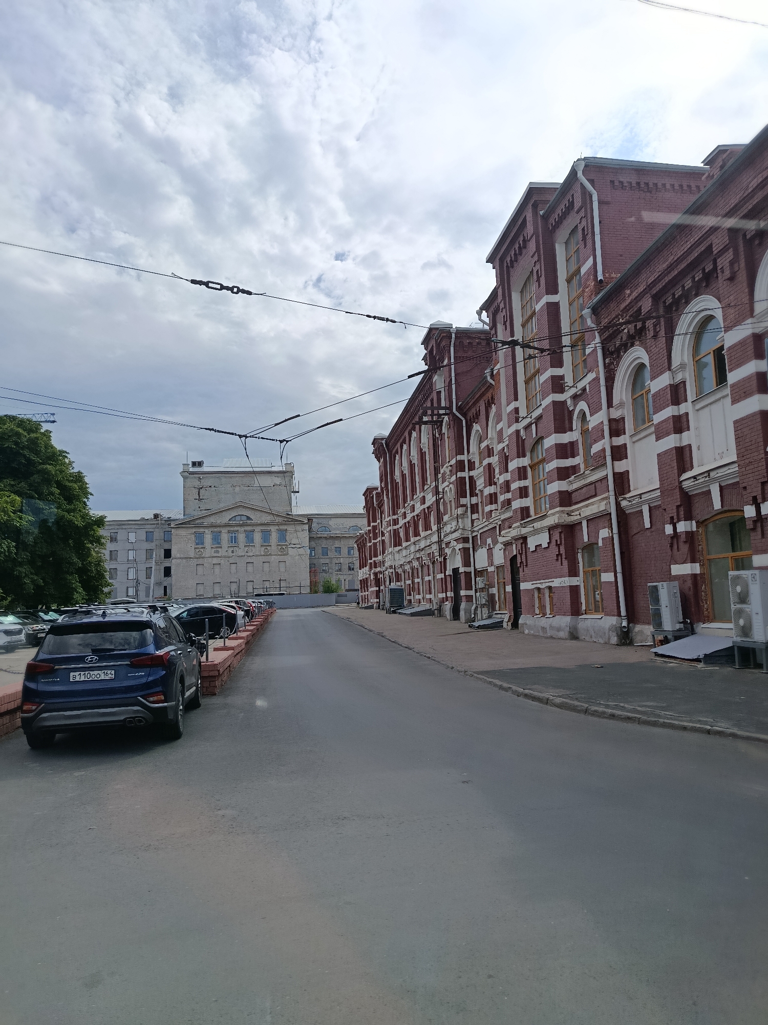 The image depicts a street scene in Saratov, Russia, with two distinct architectural styles of buildings visible. On the right side of the street, a long, imposing building constructed of dark red brick with white decorative banding dominates the view. This building features numerous windows with arched tops, some of which are yellow. The building has multiple stories and appears to be an older industrial or institutional structure.
On the left side of the street, a different building is visible, characterized by a lighter-colored facade, possibly concrete or pale brick. This structure is larger and more modern in its design, with a prominent central section that rises higher than the surrounding wings. It has a more utilitarian appearance with regularly spaced windows.
The street itself is paved and appears to be an urban road. Several cars are parked along the right curb of the street, in front of the red brick building. One car, a dark blue SUV, is prominently positioned in the foreground on the left side of the street. Overhead, complex electrical wires, possibly for trams or trolleybuses, crisscross the sky, supported by poles. The sky is overcast with gray clouds. The scene appears to be a typical streetscape in a Russian city, likely an area with a mix of historical and more recent industrial or civic architecture.