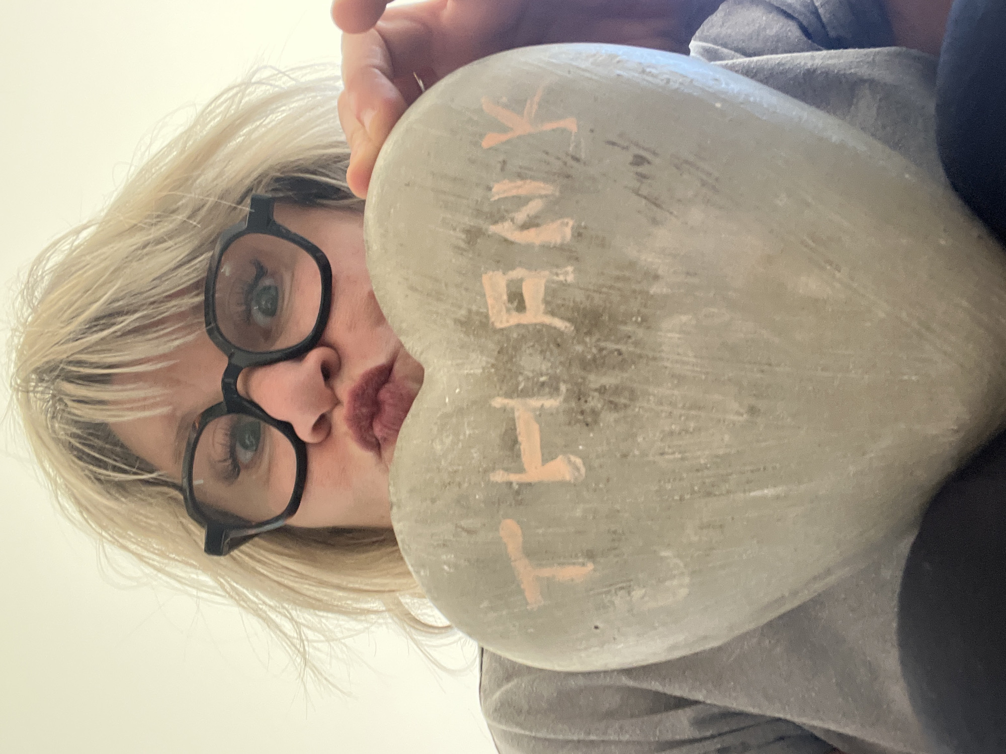 A woman with blonde hair and black-rimmed glasses is holding a heart-shaped stone up to the camera, partially obscuring her face. The stone has the word "THANK" written on it in a pale pink paint. The background is a plain white wall, and the woman is wearing a gray shirt. The location is Chambœuf, France.