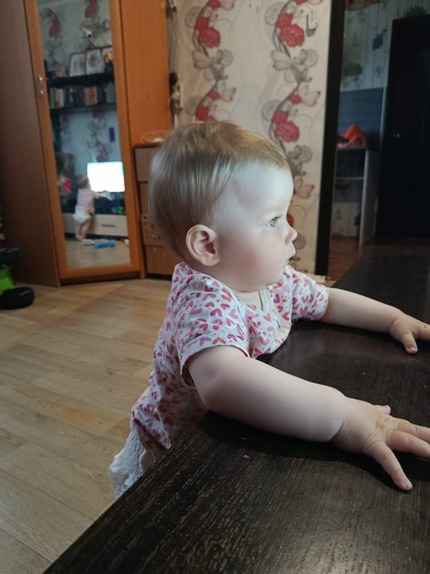 A baby wearing a white and pink heart-patterned shirt stands next to a dark brown table. The baby is looking to the right. In the background, there is a wardrobe with a mirror reflecting a room and another person. The wallpaper has a floral pattern with red flowers. The location is Фоки, Russia.