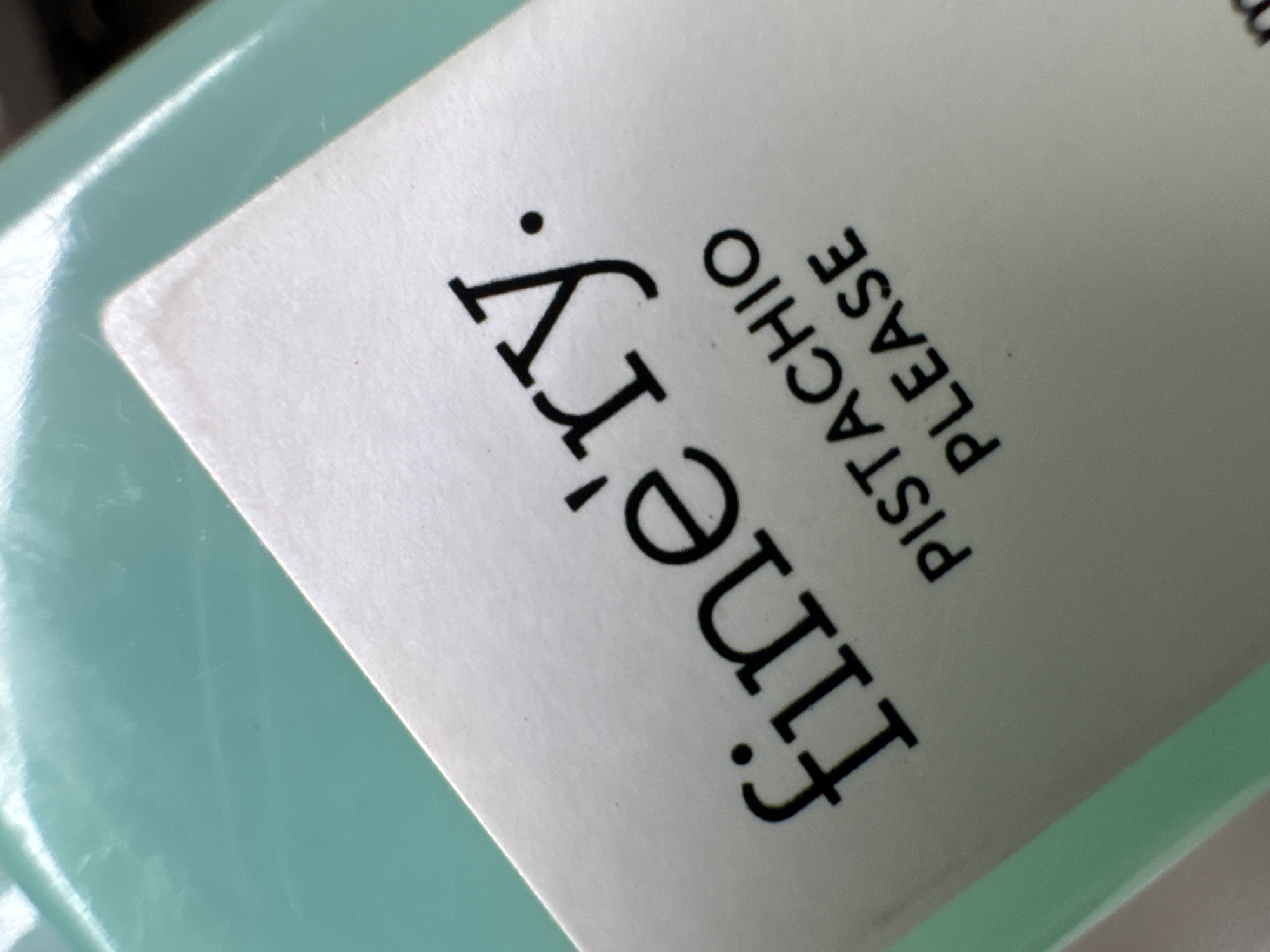 The image shows a close-up of a label adhered to a light green container. The label is white and features black sans-serif text reading "Fine'ry." vertically, followed by "PISTACHIO PLEASE" written horizontally in smaller letters. The photograph is taken at an angle, partially obscuring the full view of the container and label. There is no verifiable information in the image to indicate the location is Huixquilucan de Degollado, Mexico.
