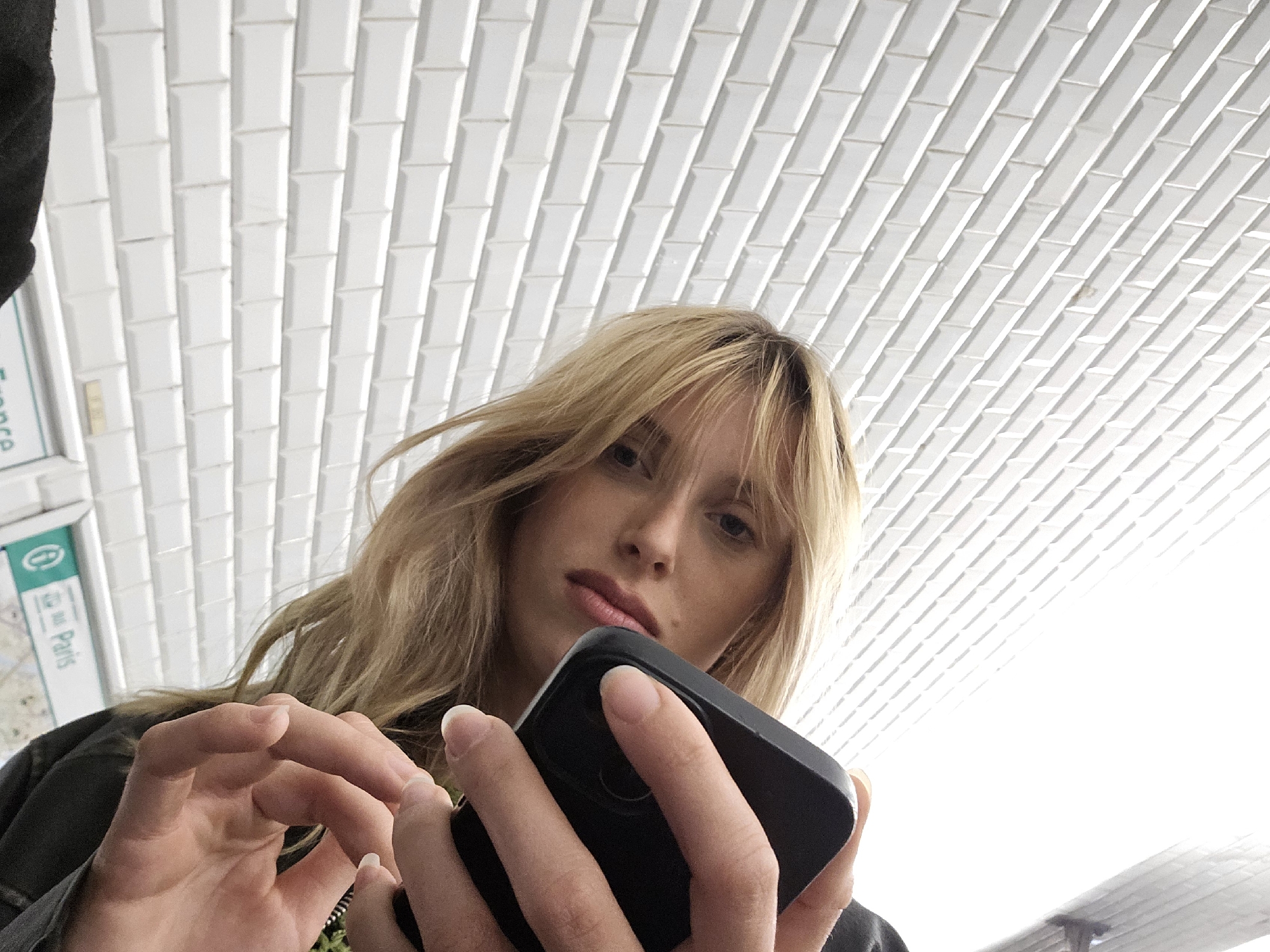 A waist-up angled shot depicts a fair-skinned young woman with blonde hair and bangs. She is holding a black smartphone in her hands, with her fingers on the screen. The woman is likely standing in the Paris metro, identifiable by the distinctive white rectangular tiles of the metro station walls behind her. A sign bearing the word "Paris" in green is visible on the left of the frame. She is wearing a black or dark-colored jacket. The environment is brightly lit, with the light source originating from above.