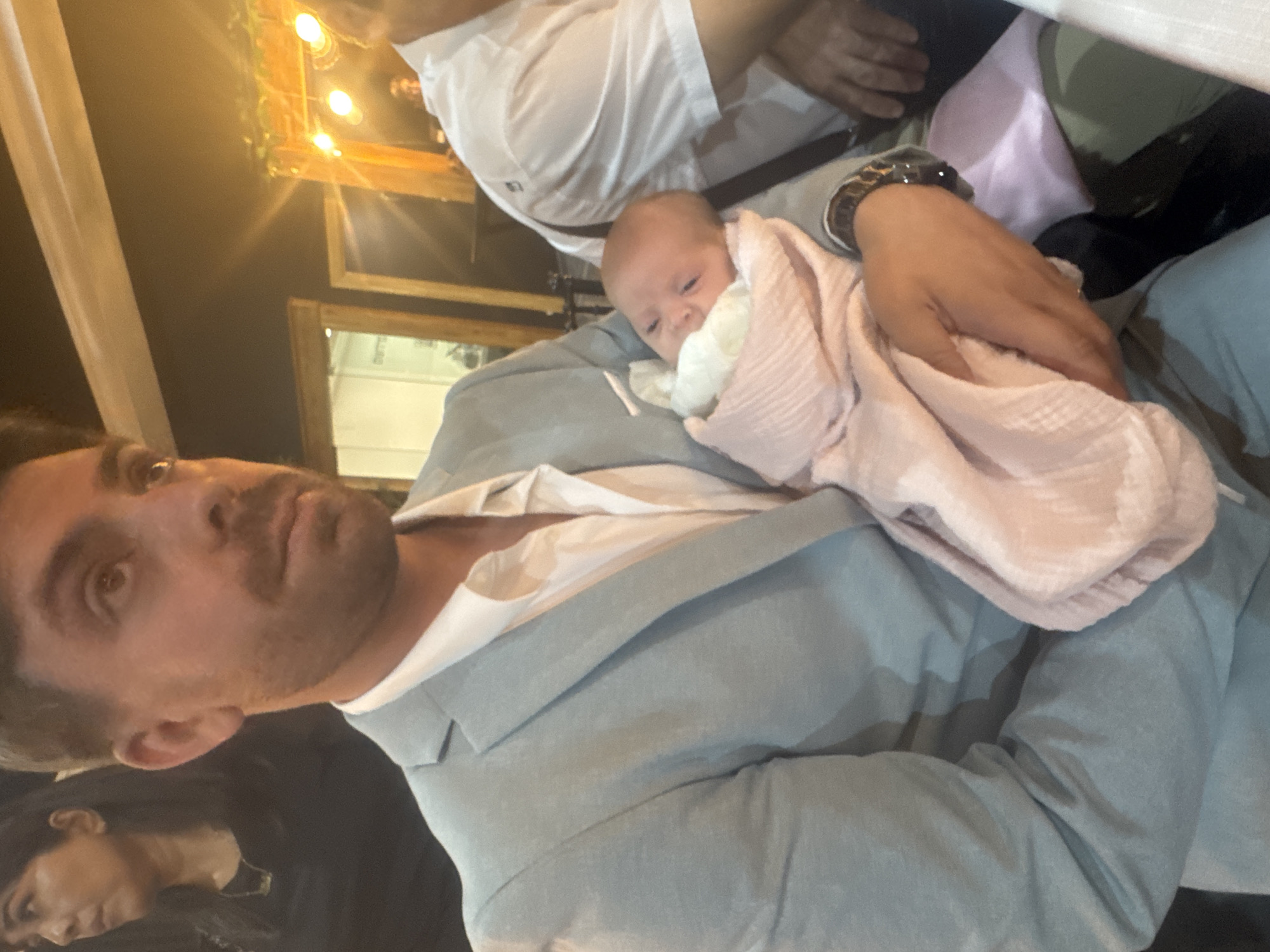 A man in a light blue suit and white shirt holds a newborn baby wrapped in a pink blanket. The man's left arm cradles the baby. In the background, a woman in a black shirt stands to the left, and another person in a white shirt is visible to the right. The environment appears to be an indoor setting, possibly a venue with decorative lighting and mirrors in the background. The image is shot from a medium close-up perspective. The location of Migdal HaEmek, Israel is not discernible from the image.