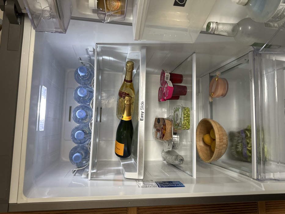 The image depicts the interior of a domestic refrigerator, open to reveal its contents. The scene is located in the United Kingdom, specifically inferred to be in London due to the presence of 'Waitrose' branded items, a major UK supermarket chain.
The refrigerator is well-lit and organized with clear shelves. On the top shelf, five clear plastic water bottles with blue caps are arranged horizontally in a metal rack. The shelf below, labeled "Easy Slide," holds two champagne bottles lying horizontally: one dark green bottle of Veuve Clicquot and one gold-foiled bottle.
The third shelf contains various packaged food items: a metallic-wrapped item to the left, a clear plastic container with dark contents, a clear plastic container of "Waitrose Garden Pea Edamame," and three red plastic pots with white lids (likely yogurt or dessert, with 'Waitrose' branding partially visible).
On the bottom shelf, a light wooden bowl filled with several yellow lemons is positioned to the left. To its right, a light pink facial roller rests on a small, round, translucent container. A green package, likely "Rosemary Salad," is partially visible directly below these items.
The right side of the image shows partial views of the refrigerator door shelves, containing a clear plastic holder with an amber-colored bottle and, on a lower shelf, a tall, clear glass bottle with a green cap next to a white plastic bottle with a blue cap. The refrigerator's exterior appears to be metallic grey, and a wooden panel is visible to the far left of the frame. No people are visible.