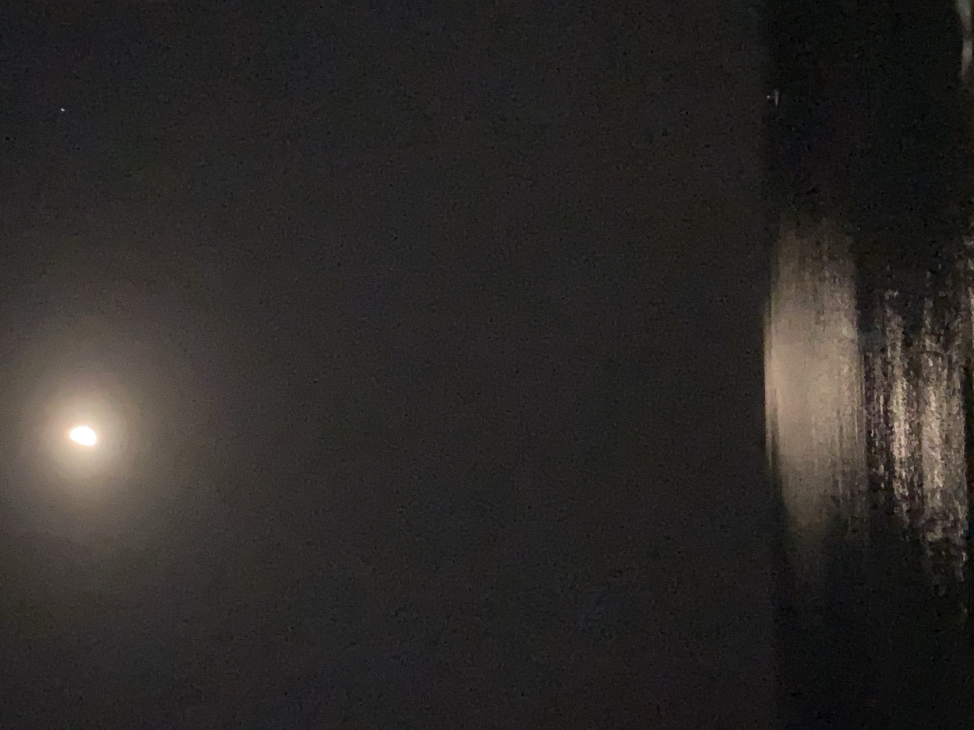 The image shows a nighttime scene of the sea, with a bright crescent moon in the upper left quadrant of the sky. A reflection of the moon shimmers on the dark water, creating a path of light. The horizon is visible in the lower part of the image, separating the sea from the sky. There appears to be a faint light in the far distance on the water, possibly a boat. The environment is dark and serene, characteristic of a moonlit night over the sea. The specific location in Alanya, Turkey, is not discernible from the visual information provided in the image.