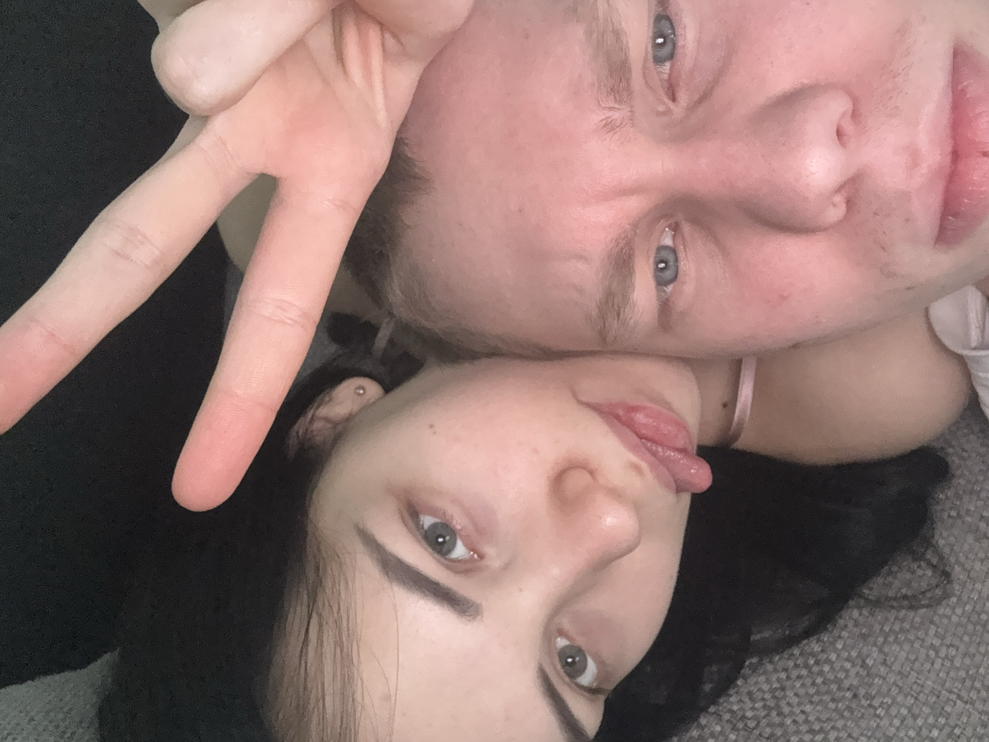 A close-up, overhead selfie shows a couple. The woman on the left has dark hair and is sticking her tongue out playfully. She has a small earring in her left ear and is wearing a pink ribbon around her neck. The man on the right has fair skin and blondish hair. His eyebrows are furrowed, and he is looking directly at the camera. Both have blue eyes. The woman is holding up two fingers in a peace sign near the top of the frame. They appear to be lying down on a grey, textured surface, possibly a couch. The background is dark and out of focus.