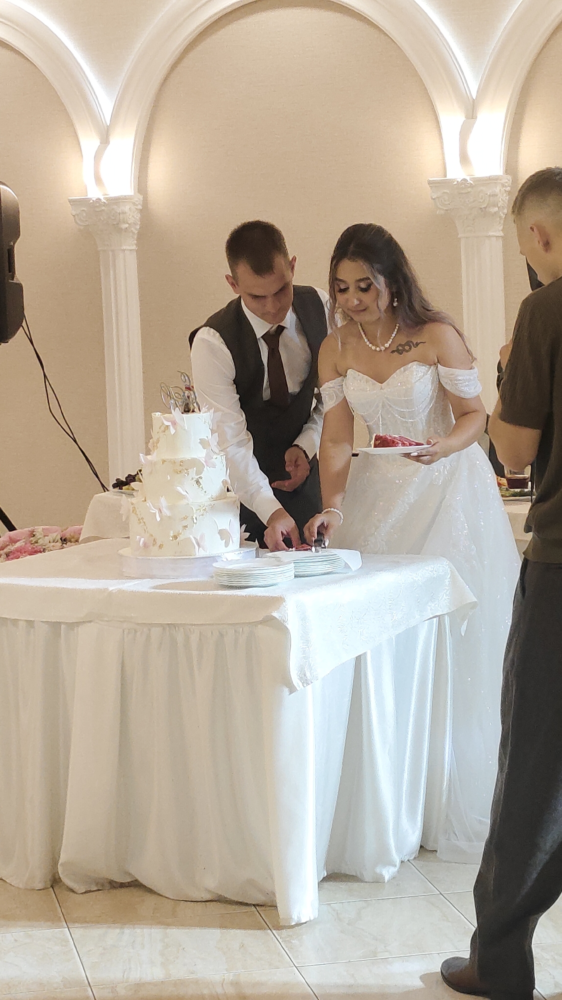 An outdoor wedding ceremony takes place in Beryozovka, Russia, with a couple cutting a tiered white wedding cake. The bride, in a white sequined off-the-shoulder wedding gown, holds a plate with cake as the groom, in a dark vest and white shirt, uses a knife to cut the cake. They are standing behind a table covered with a white tablecloth adorned with a floral arrangement. Several guests are present, including a person in a dark shirt standing to the right. The background features decorative architectural elements with soft lighting.