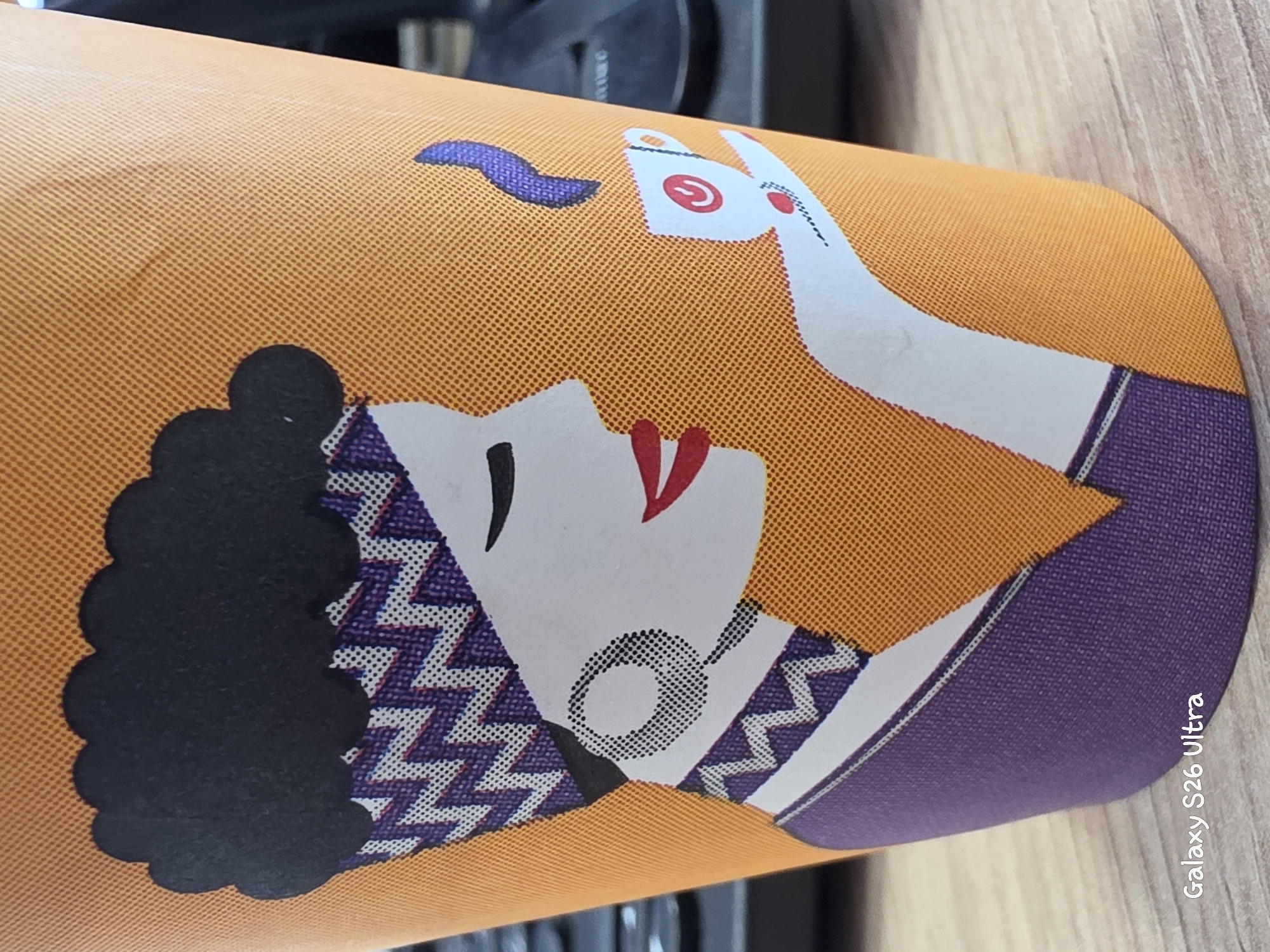 The image displays a close-up of a paper cup with an artistic design of a woman's profile. The woman has dark, voluminous hair, a patterned headscarf in shades of purple and white, and a large circular earring. She is depicted with closed eyes, red lips, and is holding a small white cup with a red symbol on it, from which a wisp of blue smoke is rising. The cup itself is orange with a textured pattern of small dots. The bottom left corner of the image shows the text "Galaxy S26 Ultra". The background is blurred but appears to be a wooden surface. The image does not contain any information about the city of Plovdiv, Bulgaria.