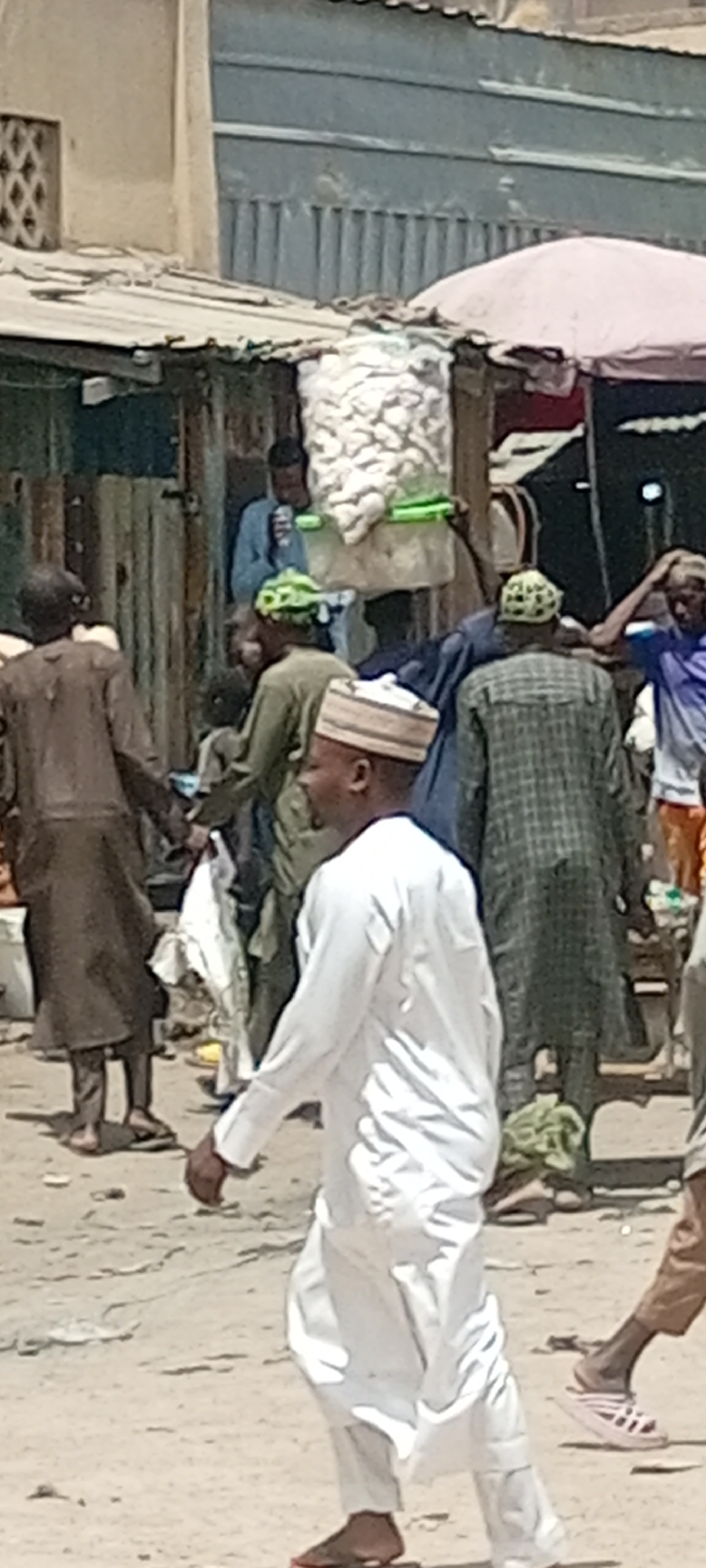 The image captures a bustling street scene in Fagge C, Nigeria. People are walking in various directions, creating a sense of movement. Some individuals are wearing traditional clothing, including robes and hats. A large white bag, possibly containing goods for sale, sits on a stand or cart. Buildings with metal siding and corrugated roofs line the street, while a light-colored umbrella offers shade. The ground is light-colored, suggesting a dirt or sandy surface.