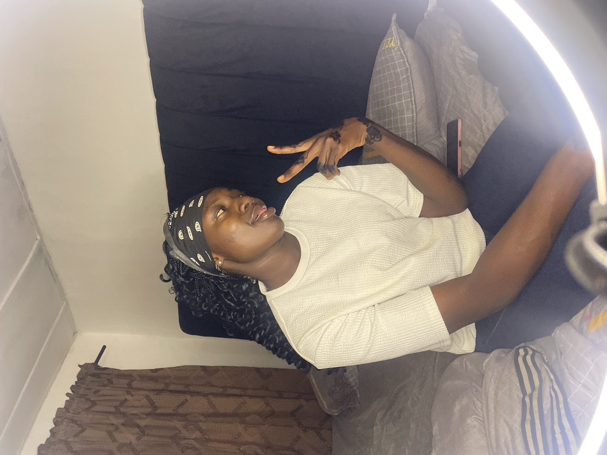A young Black woman is lying on a bed in a room. She is wearing a white shirt and a black and white patterned headscarf. Her long, black braided hair cascades behind her head. She is lying at an angle, with her head slightly turned and tilted back. Her tongue is visible. Her left hand is raised, with two fingers extended in a "peace" sign. There appears to be dark paint or henna on the back of her hand and fingers. A phone is visible on the bed beside her. A ring light is positioned near the camera, creating a bright glow around the edges of the frame.
The bed has several pillows and a black headboard. Part of a brown curtain can be seen in the lower left corner of the frame. The room has white walls.
There is no definitive information in the image to confirm that the location is in the city of Jos, Nigeria.