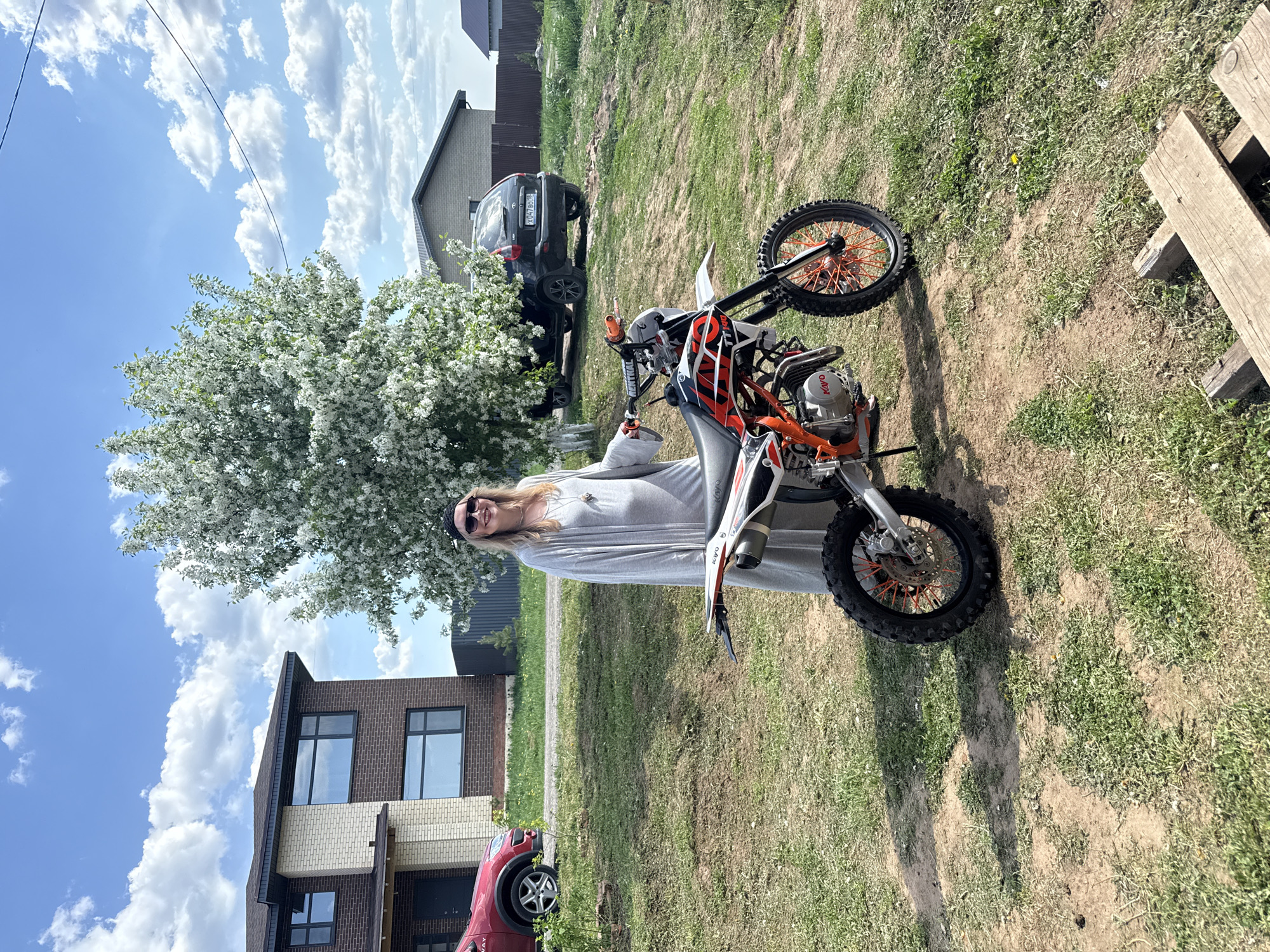 A woman in sunglasses and a gray dress is standing next to a dirt bike on a grassy lawn. A house with brick accents and a red car parked in front is visible to the left, and a black car and another house are in the background. A flowering tree is beside the woman. The sky is blue with scattered white clouds. The location appears to be Izhevsk, Russia.