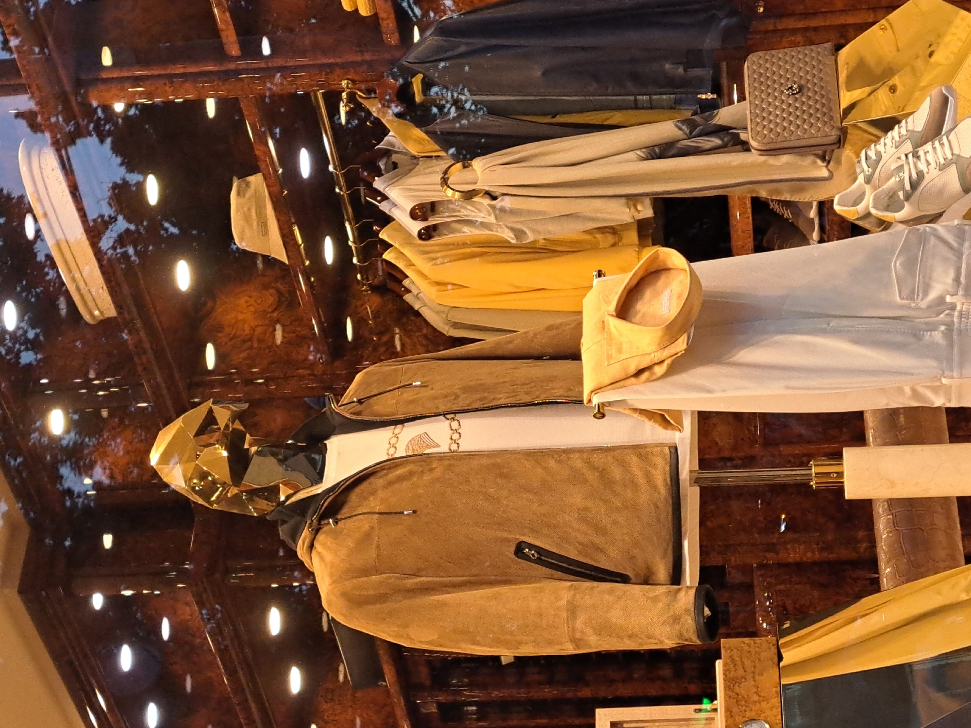 The image shows a clothing store display in Yerevan, Armenia, featuring a mannequin with a gold geometric mask wearing a brown suede jacket over a white shirt. A rack of clothing behind the mannequin includes blue, beige, and yellow items. A clutch bag and sneakers sit on top of the garments. The store window reflects the outside environment and decorative lights.