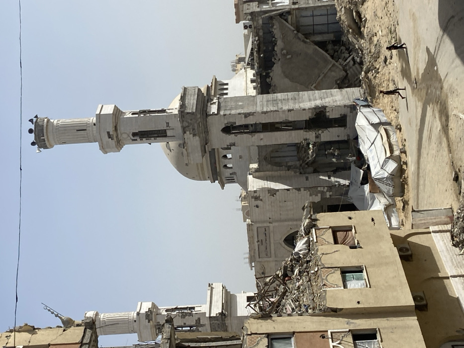 The image depicts a devastated urban landscape in Khan Yunis, Palestinian Territory, under a clear, light blue sky.
In the background, a large mosque complex dominates the scene. It features a central, light-colored dome and two minarets. The minaret on the right is tall and cylindrical, topped with multiple loudspeakers, and exhibits significant structural damage, including large cracks and missing sections along its shaft. The left minaret, partially obscured by a damaged building, also shows damage at its finial. The mosque's walls are extensively damaged, with large sections of material missing, exposed rebar, shattered arched windows, and widespread pockmarks. Arabic script, reading "مدرسة الأستاذ محمد على سالم" (School of Professor Muhammad Ali Salem), is visible on a facade section between the minarets.
In the left foreground, a multi-story building is severely damaged. Its upper floor has partially collapsed, leaving a heap of rubble, twisted rebar, and debris on its roof. The remaining walls are pockmarked, and windows are broken or missing. To the right of this building, closer to the mosque, a large section of another structure has completely collapsed, forming a substantial mound of concrete and metal debris. Adjacent to this rubble, several makeshift shelters constructed from white tarpaulins are visible on the sandy ground.
The ground in the immediate foreground and extending into the midground is a mix of sand, dirt, and construction rubble. A dark, possibly paved, road or path traverses the lower portion of the image horizontally, showing some dark stains or patches. Two individuals, appearing as children or young adults, dressed in dark clothing, walk barefoot along this path in the lower-right quadrant, casting shadows to their right. A thin power line or cable stretches across the top of the frame.