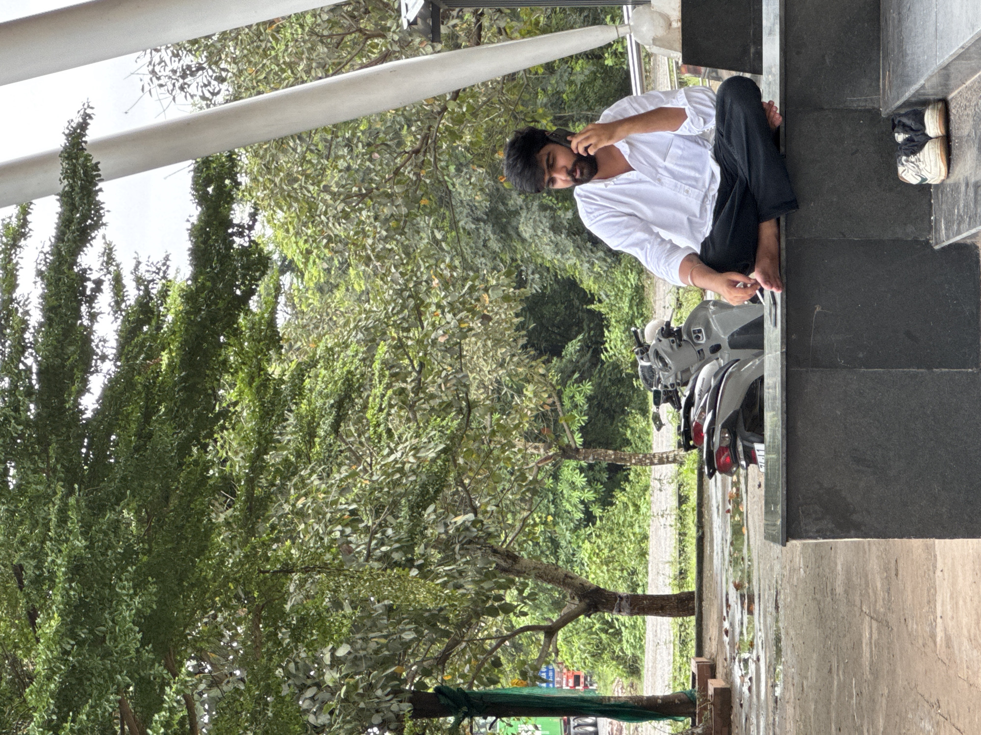 A young man with dark hair and a beard, wearing a white button-down shirt and black pants, is sitting cross-legged on a dark stone ledge, talking on his mobile phone. His bare feet are visible, and a cigarette is held between his fingers. Next to him on the ledge is a smartphone. In front of the ledge, on the ground, are a pair of white and black sneakers. To the left of the man, a row of scooters is parked. The environment is outdoors, with lush green trees and foliage forming the background. The ground appears wet, with puddles reflecting the surrounding environment, suggesting recent rainfall. In the far background, a truck is partially visible. The scene is set in Lao Bảo Commune, Vietnam, characterized by the abundant tropical vegetation and the presence of vehicles like scooters and trucks typical of the region. The overall atmosphere is casual and unhurried, despite the presence of technology and vehicles.