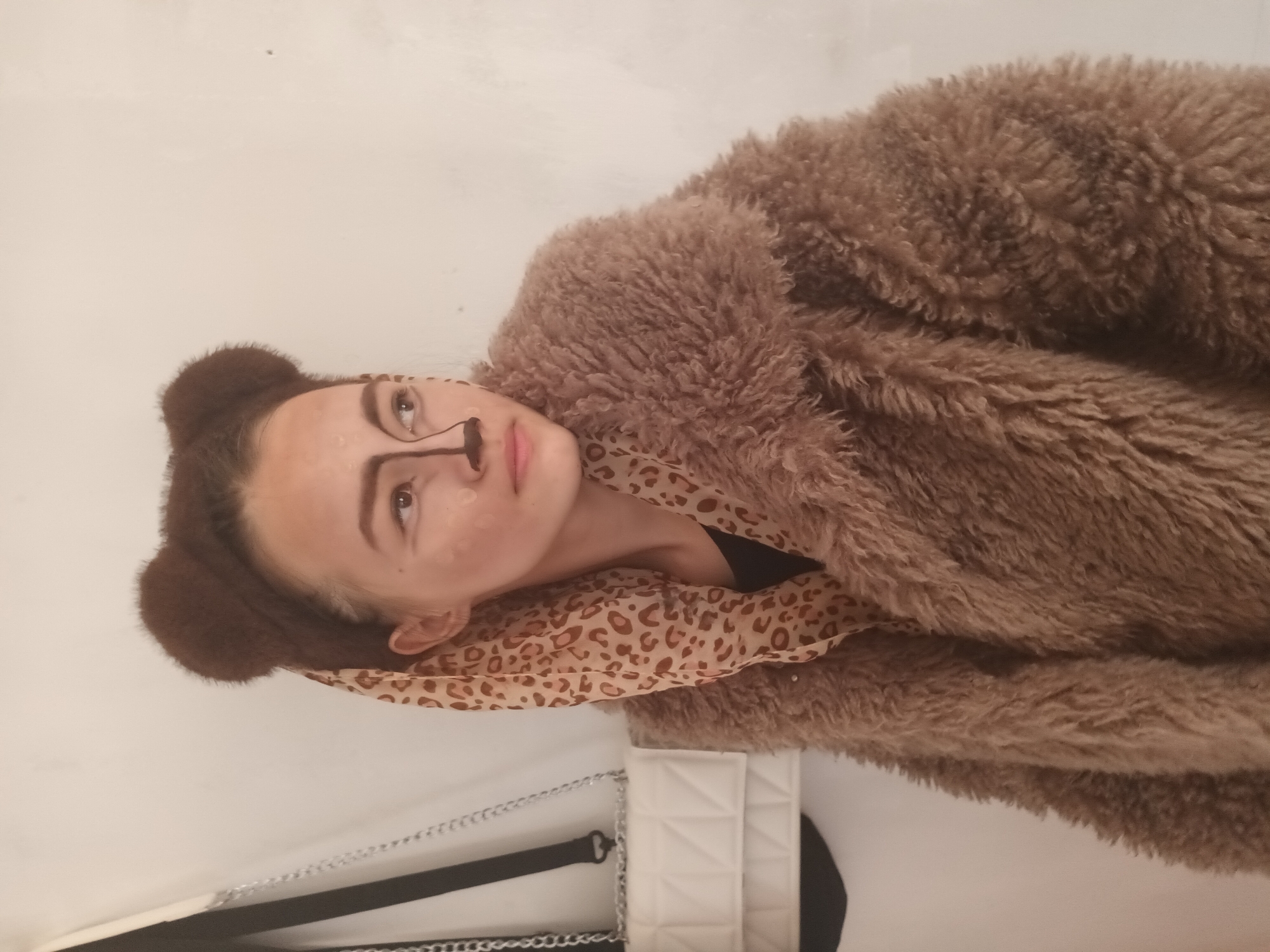 A person dressed as a bear is photographed against a white background. They are wearing a brown, furry coat and have brown bear ears attached to their head. Their face is partially covered in makeup to resemble a bear, with a black line drawn from their nose to their upper lip. They are wearing a patterned scarf around their neck. A white bag with a black strap and silver chain is visible in the lower left corner of the frame. No information about the location in Ольховка, Russia can be determined from the image.