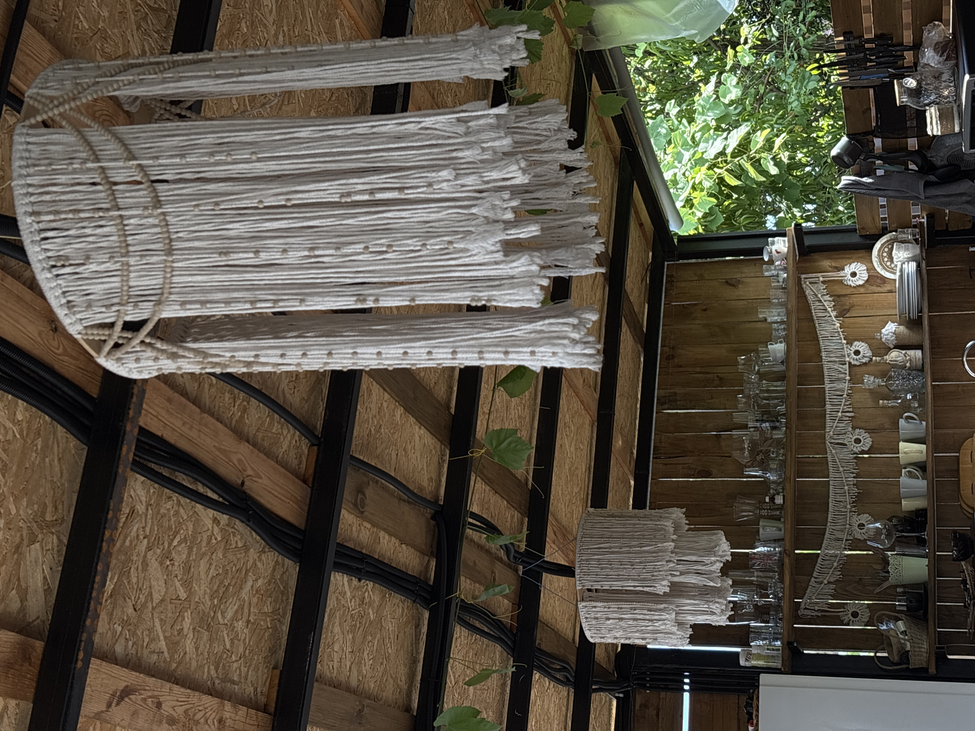 The image depicts an outdoor covered area, likely a veranda or patio, with a wooden ceiling supported by dark metal beams. Two macrame chandeliers hang from the ceiling. The larger chandelier, in the foreground, is made of white rope with beads, featuring multiple tiers of fringe. A smaller, cylindrical macrame chandelier is visible in the background.
Below the chandeliers, wooden shelves are mounted on a vertical wooden wall. The upper shelf holds a collection of clear glass bottles and various drinking glasses. The middle shelf displays a macrame wall hanging with circular, floral-like decorations, and below it, plates are stacked. On the bottom shelf, various kitchen items are visible, including cups, bottles, and a woven basket.
To the left, a white refrigerator is partially visible. To the right, lush green foliage from trees and vines can be seen, suggesting a natural, possibly garden, setting. Black electrical conduits are routed along the ceiling beams. The scene suggests a relaxed, possibly rustic, outdoor living or dining space in Gudauta, Abkhazia, given the provided context. No people are present in the image.