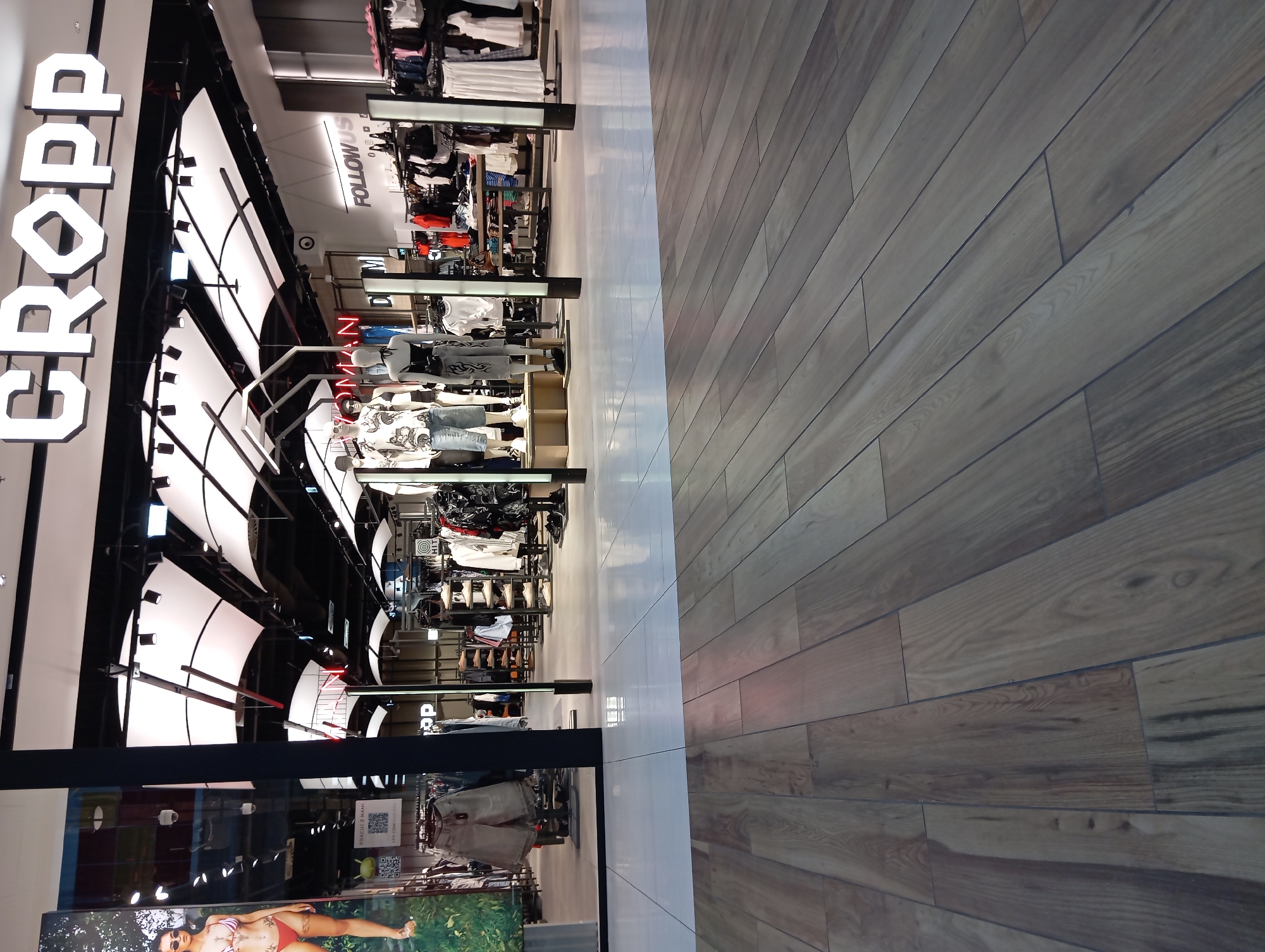 The image shows the exterior of a CROPP clothing store in the city of Wołomin, Poland, located in a commercial area or mall. The storefront features a large, illuminated "CROPP" logo. Inside, mannequins are displaying clothing, and racks of garments are visible, suggesting a retail environment. The floor in the foreground is made of wood-like tiles, and the area behind it is tiled with lighter-colored tiles. There are no identifiable people in the visible portion of the scene.