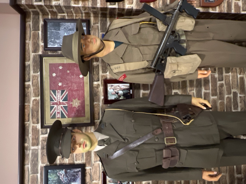 Two mannequins dressed in military uniforms are displayed against a brick wall adorned with framed images, including one of the Australian flag. The upper mannequin wears a brimmed hat and carries a firearm. The lower mannequin also wears a military hat and is equipped with a belt and holster. The scene is likely part of a museum or historical exhibit.