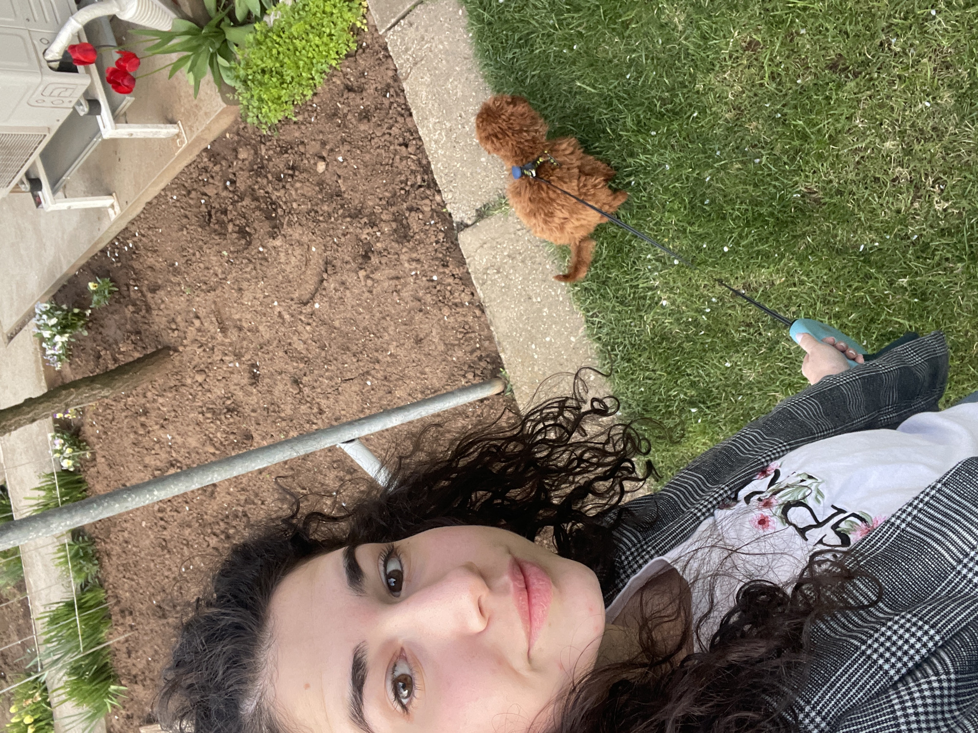 A selfie-style photograph shows a woman holding a dog on a leash. The woman has dark, curly hair and is wearing a grey and white checkered blazer over a white t-shirt. Her face is centered in the foreground. Behind her is a garden setting, including soil, grass, flowers, and a concrete walkway. A small brown poodle is standing on the grass next to the walkway, attached to a leash held by the woman. The location is presumed to be Sofia, Bulgaria, based on the prompt.