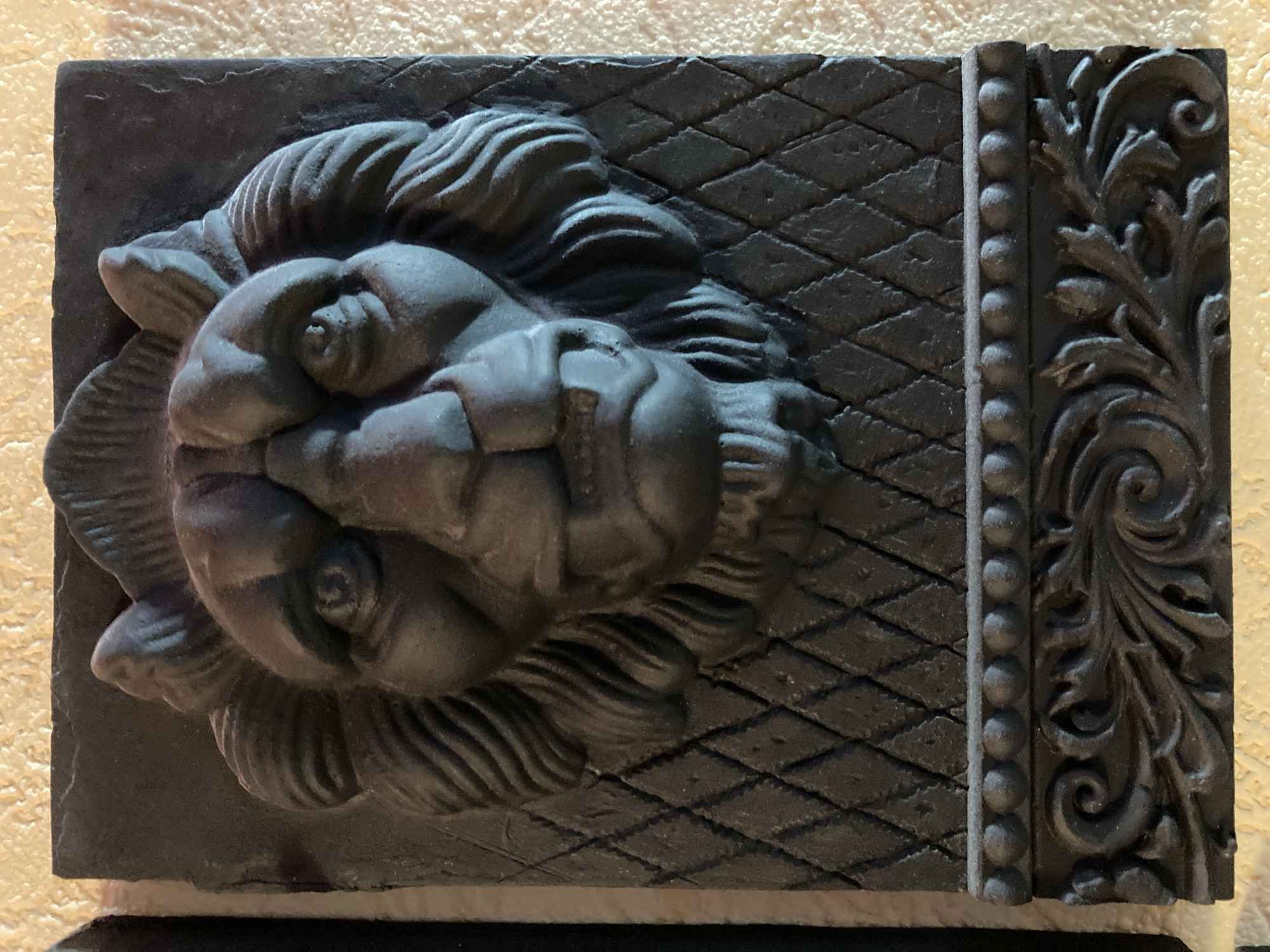 A dark gray, relief sculpture of a lion's head is mounted on a textured, light beige wall. The lion's head is centrally positioned and appears to be made of stone or a similar material. It has a fierce expression with prominent eyes, a wide nose, and an open mouth showing teeth. The lion's mane is depicted with textured strands radiating outwards. Below the lion's head, there is a textured pattern of diamonds. Further down, a decorative border of small, raised circles sits above an ornate, carved pattern resembling foliage and swirls. The lighting in the image casts shadows that highlight the depth and texture of the sculpture. The location is listed as Unknown, Unknown.