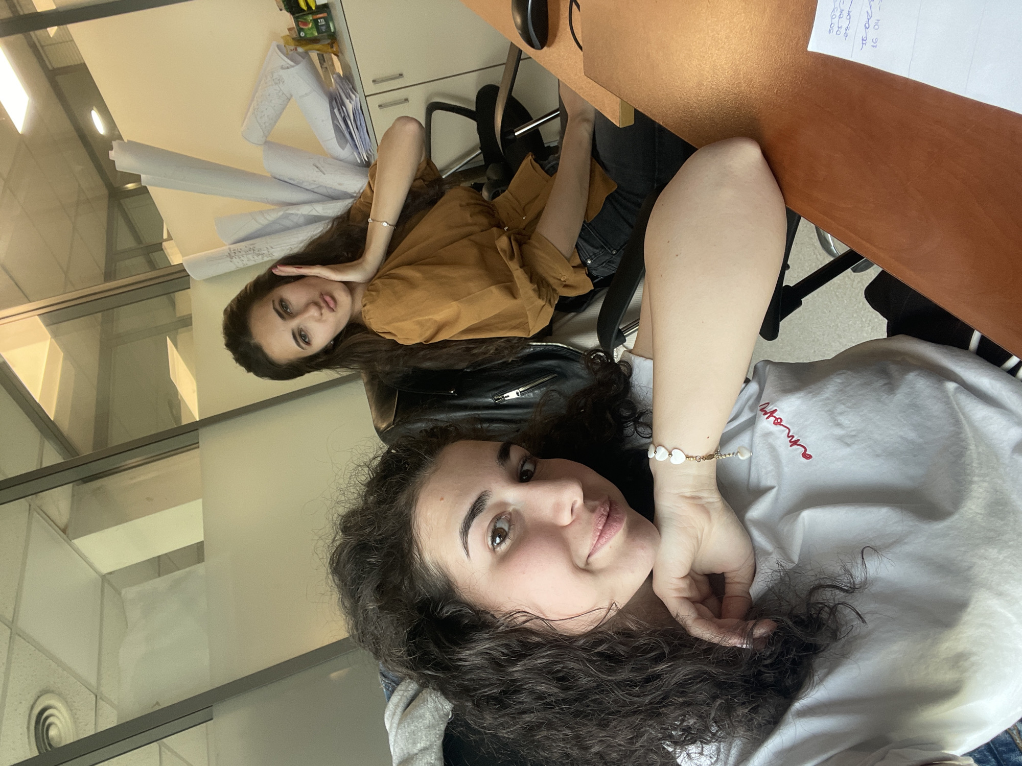Two young women are captured in a casual, posed photograph within what appears to be an office or workspace in Sofia, Bulgaria. The image is framed with the subjects seemingly seated, looking directly at the camera.
In the foreground, a woman with dark, curly hair rests her left hand near her chin, leaning slightly into the frame. She wears a light-colored (possibly white or light grey) short-sleeved top with red script text "amor" visible on the chest. A delicate bracelet adorned with small, white, heart-shaped beads is on her left wrist. Her expression is relaxed, with a slight smile.
Behind her and slightly to the left, a second woman with long, dark, wavy hair is visible. She wears a brown or mustard-colored long-sleeved button-up shirt and blue jeans. Her left hand is raised near her face, possibly touching her hair or cheek. She also gazes towards the camera.
The setting suggests an office environment, featuring desks and office chairs. Behind the second woman, several rolled-up papers, resembling architectural blueprints or large-format documents, are stacked on a surface. On a wooden desk surface to the right of the foreground woman, a black computer mouse is visible, along with a piece of paper displaying some handwritten, largely illegible notes or numbers, possibly including "10-00", "12-00", "04", and "97". The background includes glass partitions and overhead lighting, indicating an indoor, commercial building. The overall atmosphere is bright and informal.